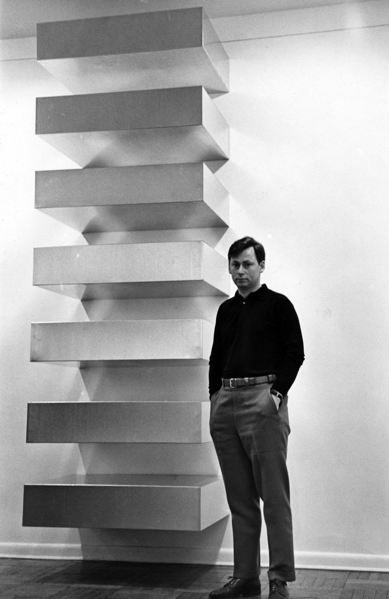 Mann steht neben einer rechteckigen Skulptur aus gestapelten Tafeln in einem minimalistischen Raum. Schwarzweißaufnahme.
