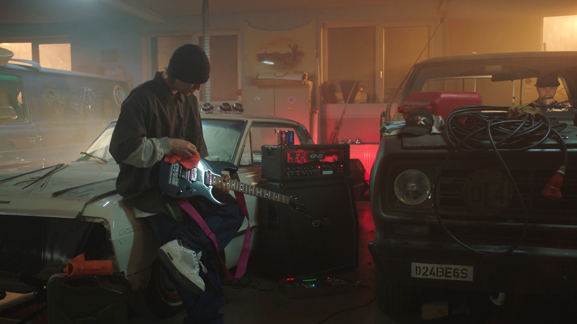 Young musician plays electric guitar in a workshop, surrounded by cars, tools, and soft lighting.
