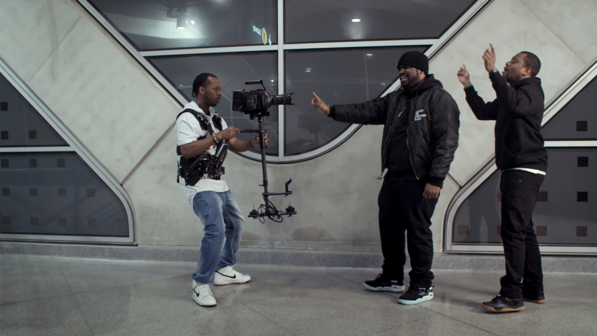 Zwei Männer posieren in einer modernen Umgebung vor der Kamera, während ein dritter Mann sie filmt.