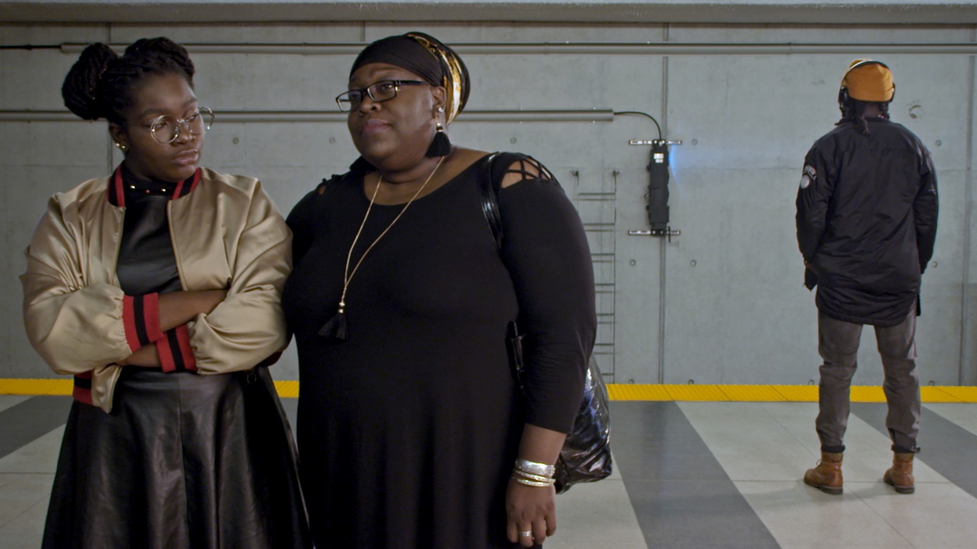 Zwei Frauen in modischer Kleidung stehen vor einem U-Bahnhof, während eine Person im Hintergrund wartet.