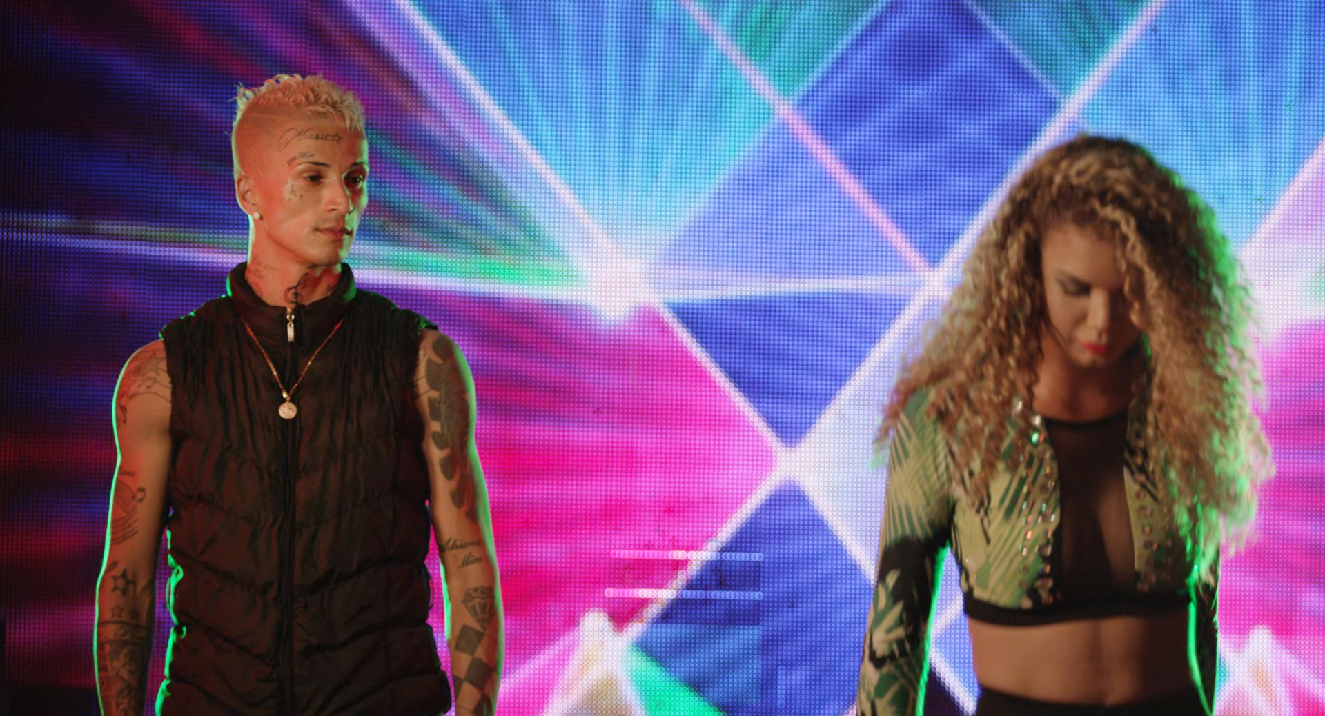 Zwei schick gekleidete Personen stehen vor einem bunten, leuchtenden Hintergrund mit geometrischen Mustern.