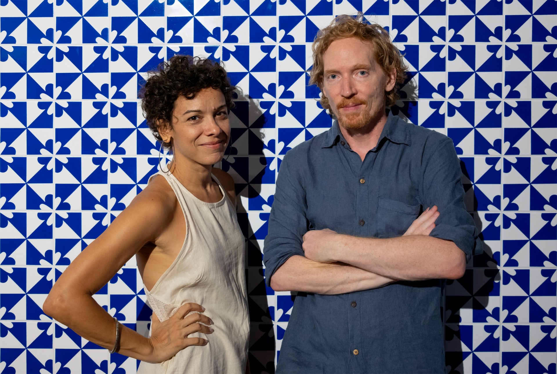 Ein Paar posiert lächelnd vor einer blauen, geometrisch gefliesten Wand in lässiger Kleidung.