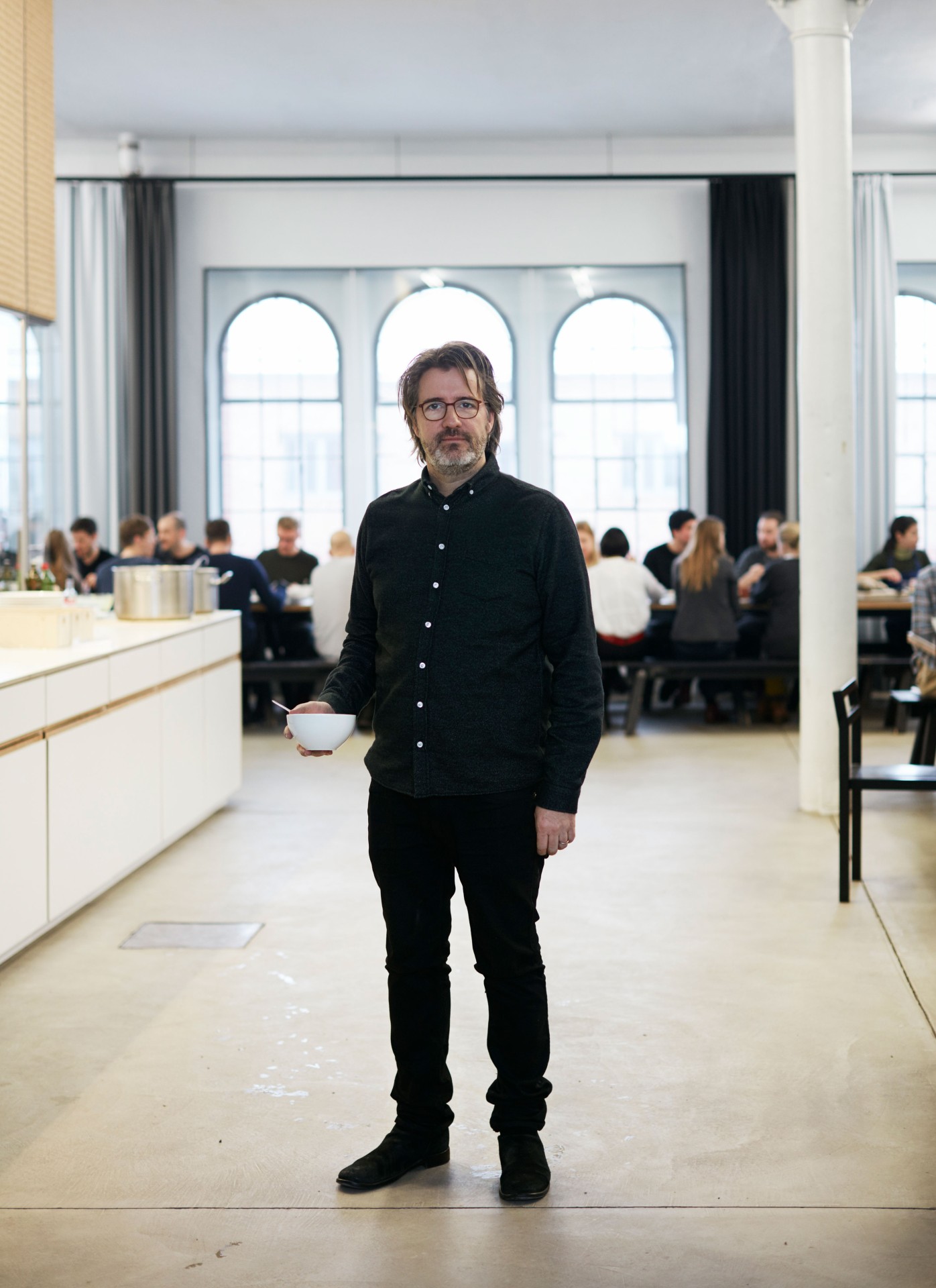 Mann mit Brille steht in einem Raum, hält eine Schüssel und schaut in die Kamera, während im Hintergrund weitere Personen sitzen.