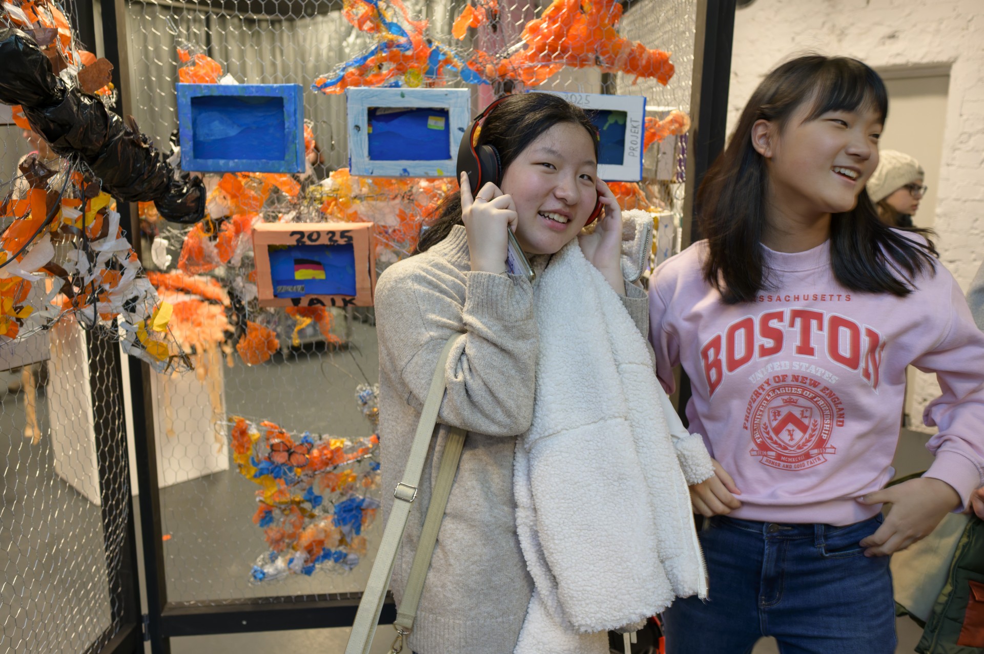 Zwei junge Frauen lächeln vor einer Kunstinstallation aus recyceltem Material. Eine von ihnen trägt Kopfhörer.