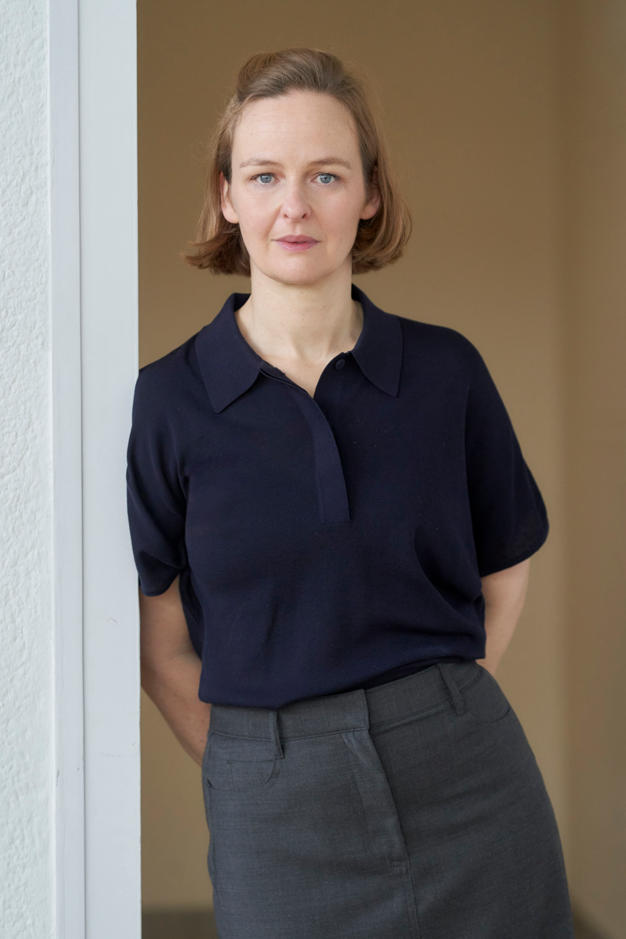 Porträt einer lächelnden Frau mit kurzem, blondem Haar in eleganter Kleidung vor neutralem Hintergrund.
