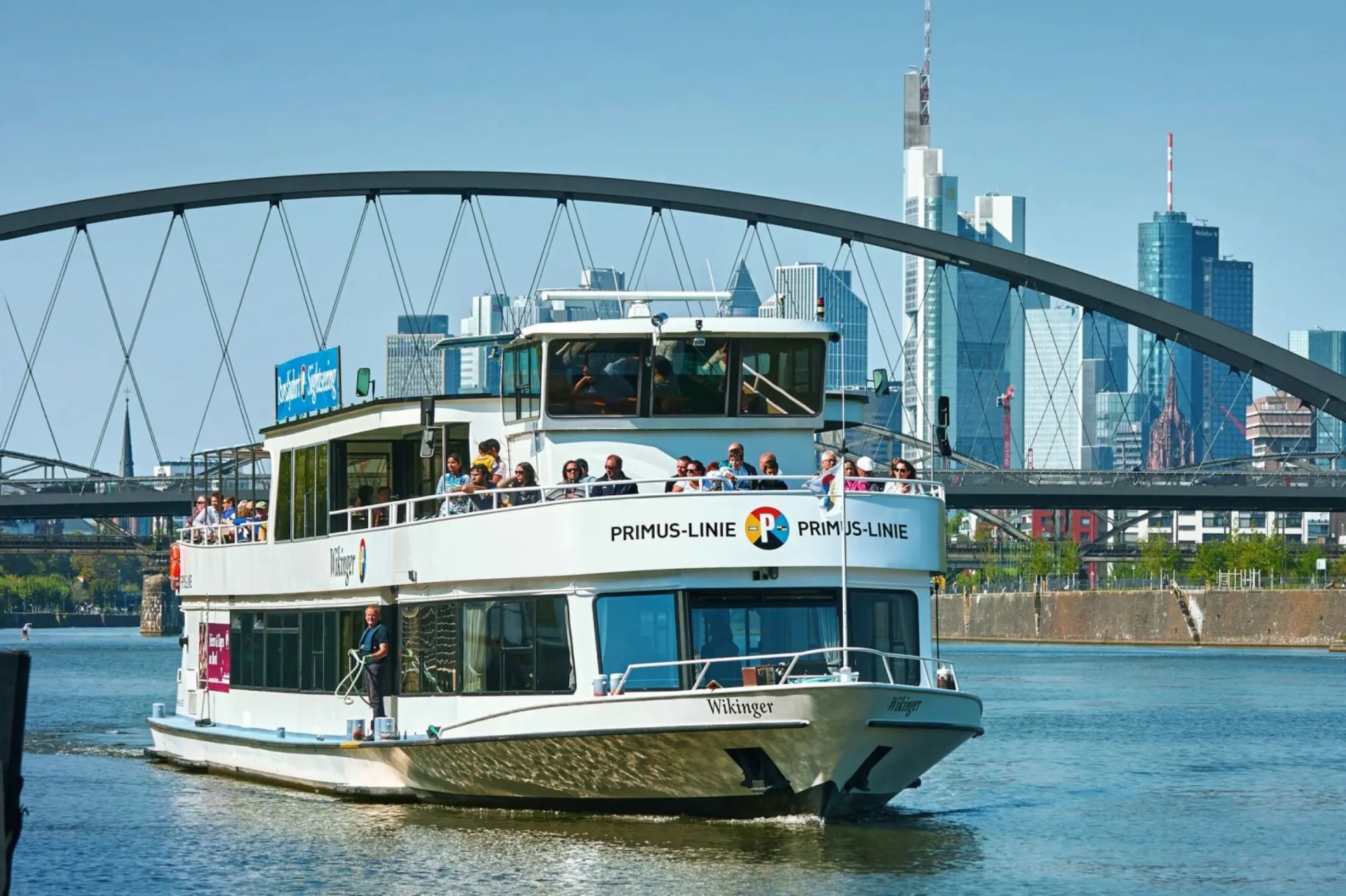 Weißes Passagierschiff auf dem Wasser, im Hintergrund eine moderne Stadt mit Brücke und Wolkenkratzern.