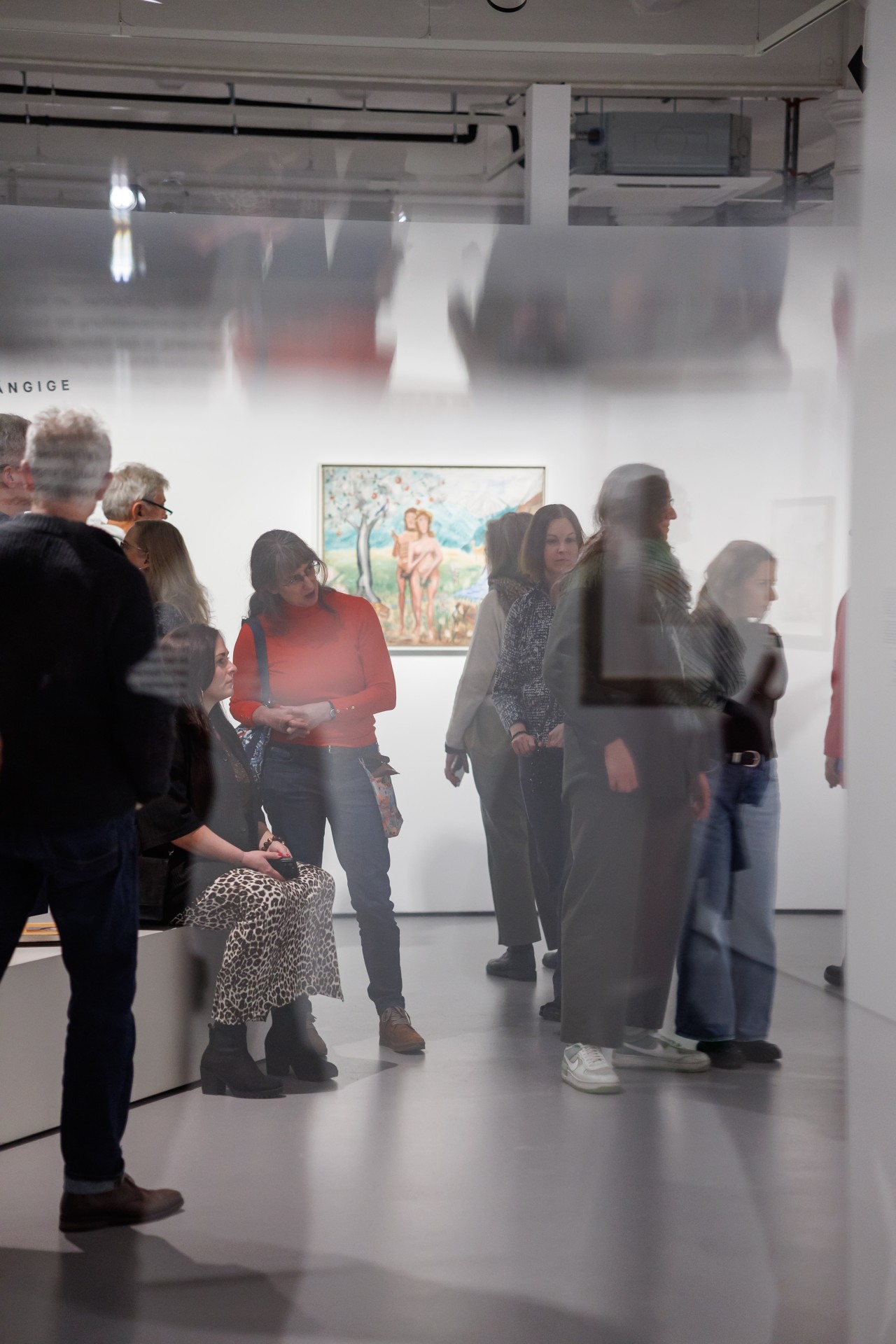 Besucher in einer Galerie betrachten Kunstwerke, während jemand sitzt und ein anderes Gespräch führt.