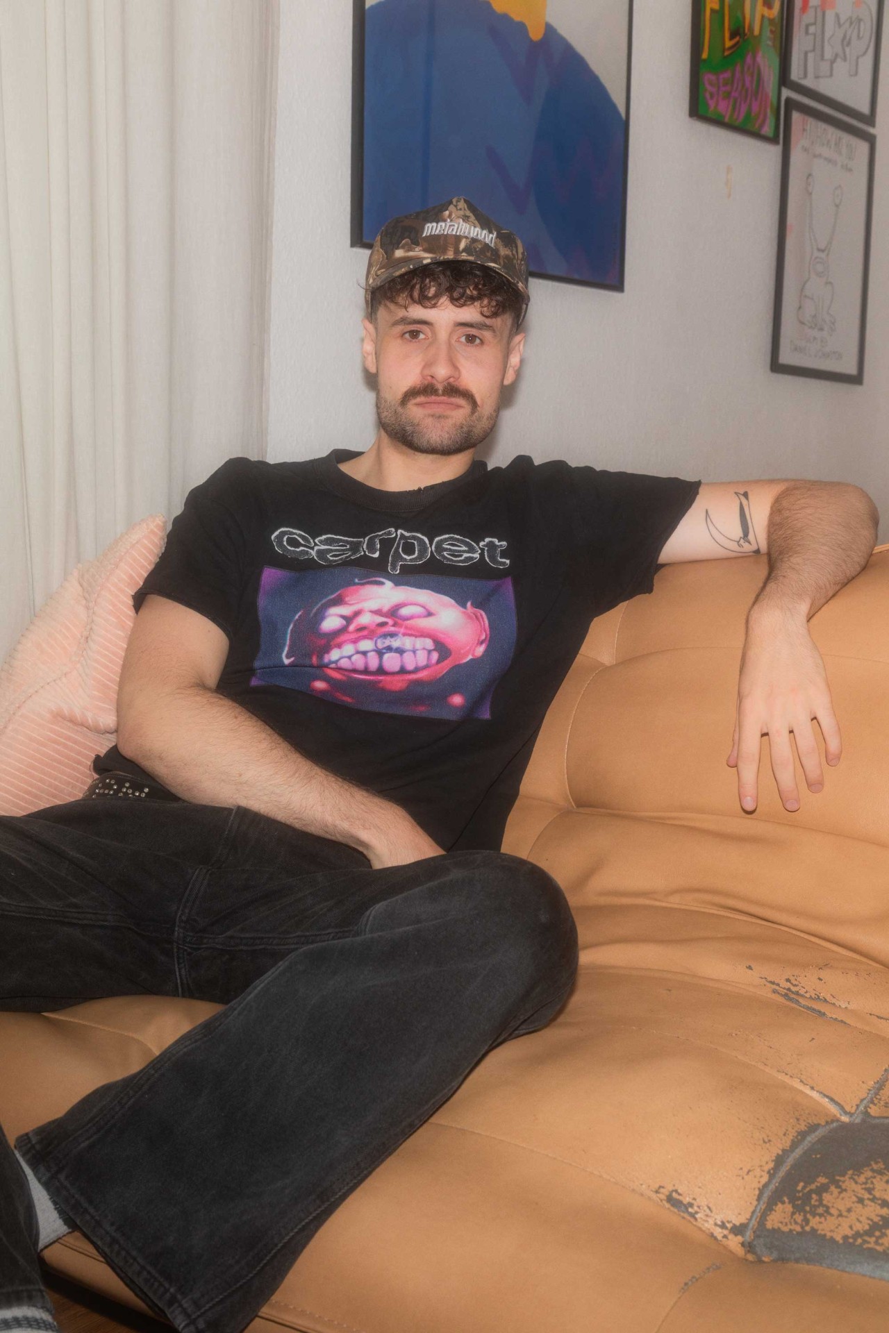 Junger Mann mit Bart in großem T-Shirt und Kapuze sitzt entspannt auf der Couch, im Hintergrund sind Kunstwerke.