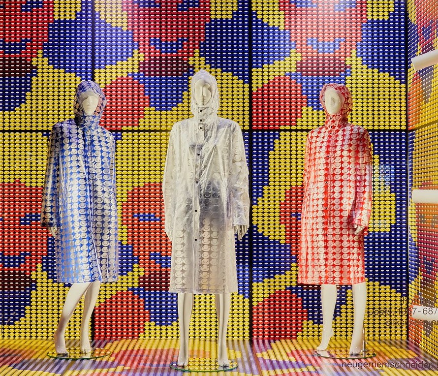 Schaufenster mit drei glitzernden Regenmänteln vor einem bunten, grafischen Hintergrund.