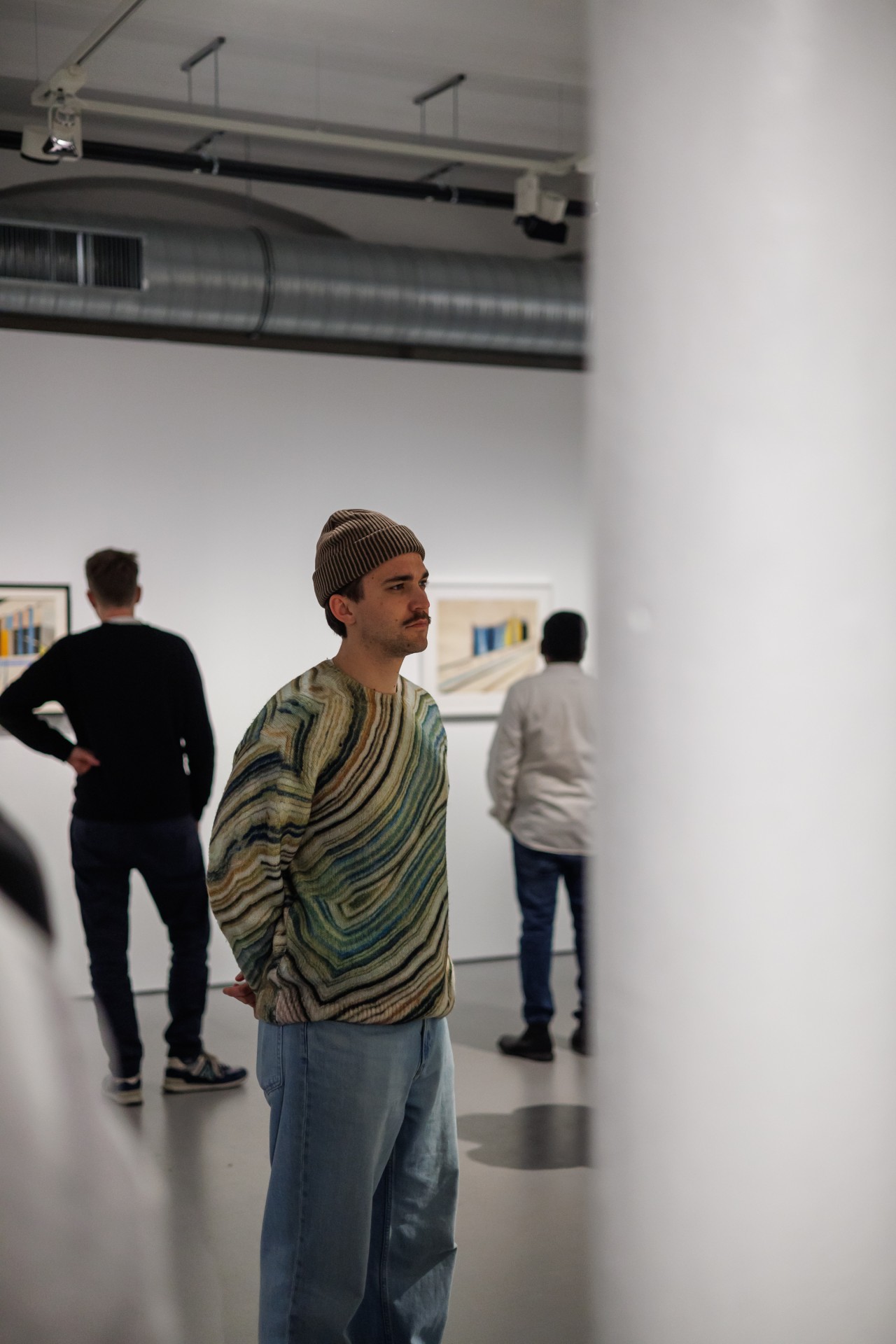 Mann in gestreiftem Pullover und Mütze betrachtet Kunstwerke in einer Galerie, während andere Besucher im Hintergrund stehen.