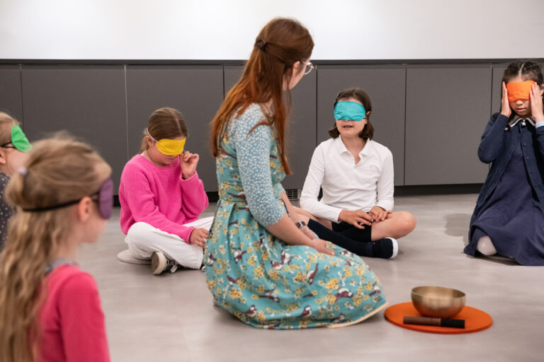 Kinder mit Augenbinden sitzen im Kreis, während eine Lehrerin ihnen etwas erklärt. Ein Klangschalen-Set ist zu sehen.