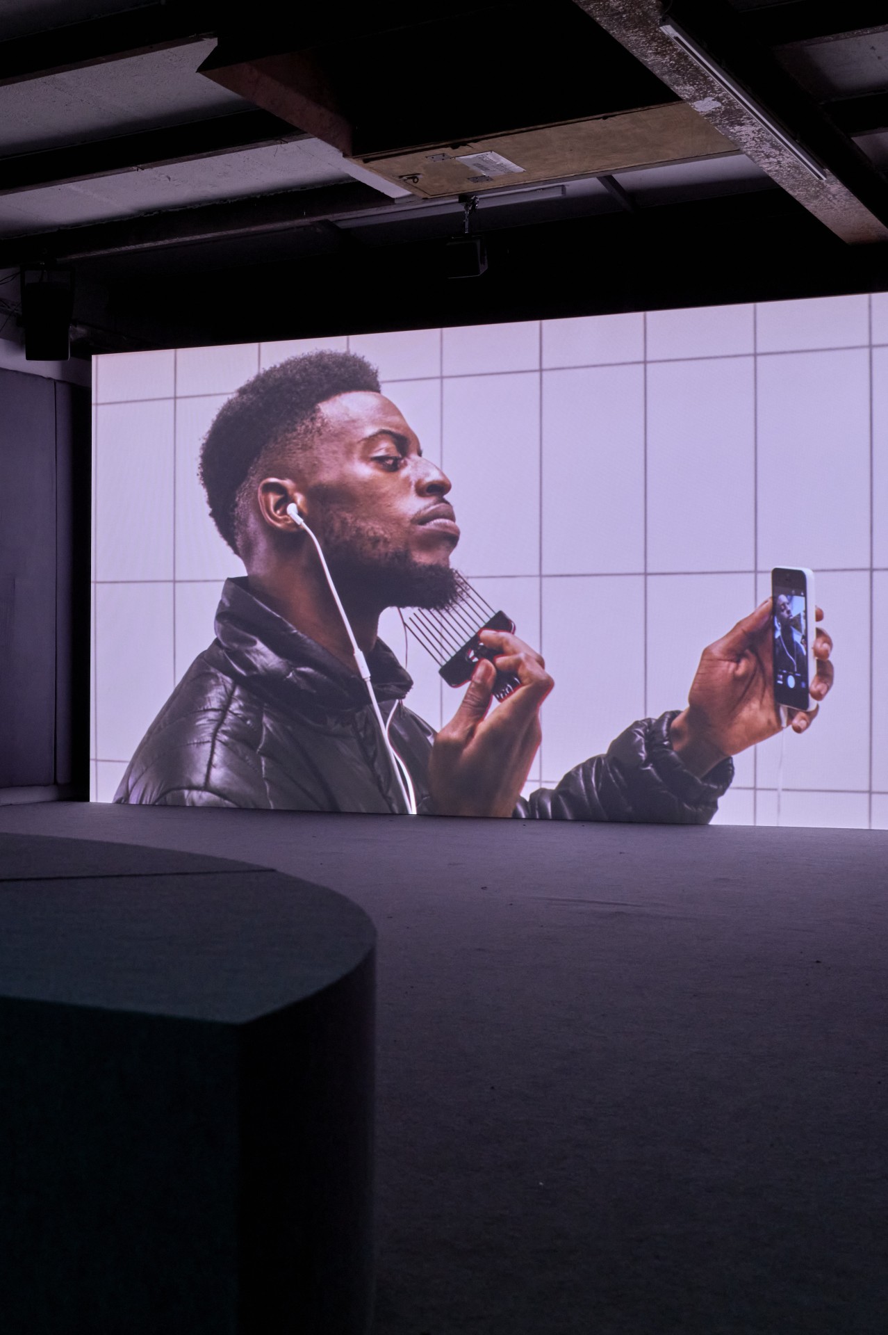 Mann mit Kopfhörern und Kamm in der Hand schaut auf sein Smartphone vor einem großen Bildschirm.