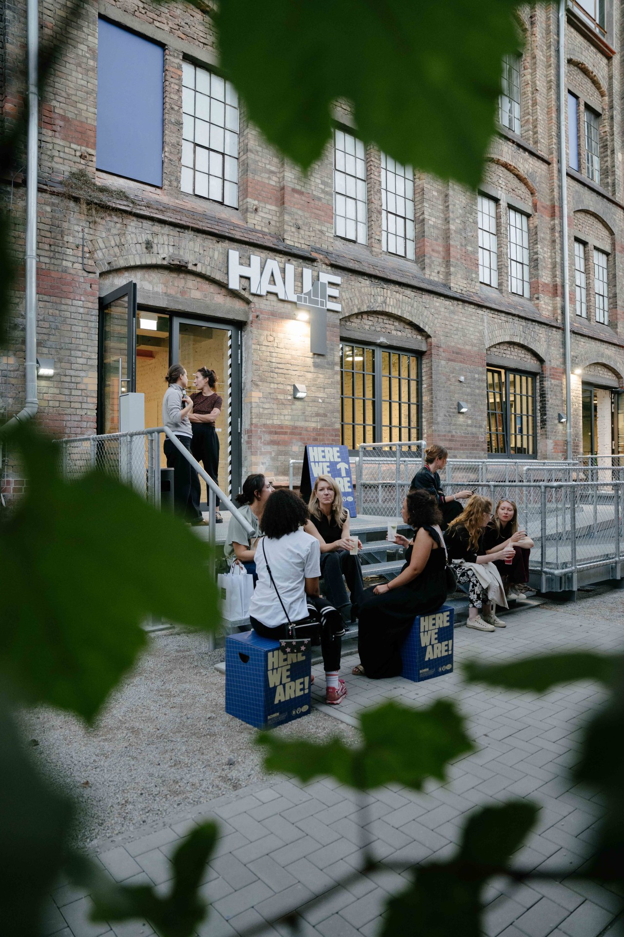 Gruppengespräche vor einer alten Fabrikhalle, umgeben von Pflanzen, mit Schildern, die "HIER SIND WIR!" zeigen.