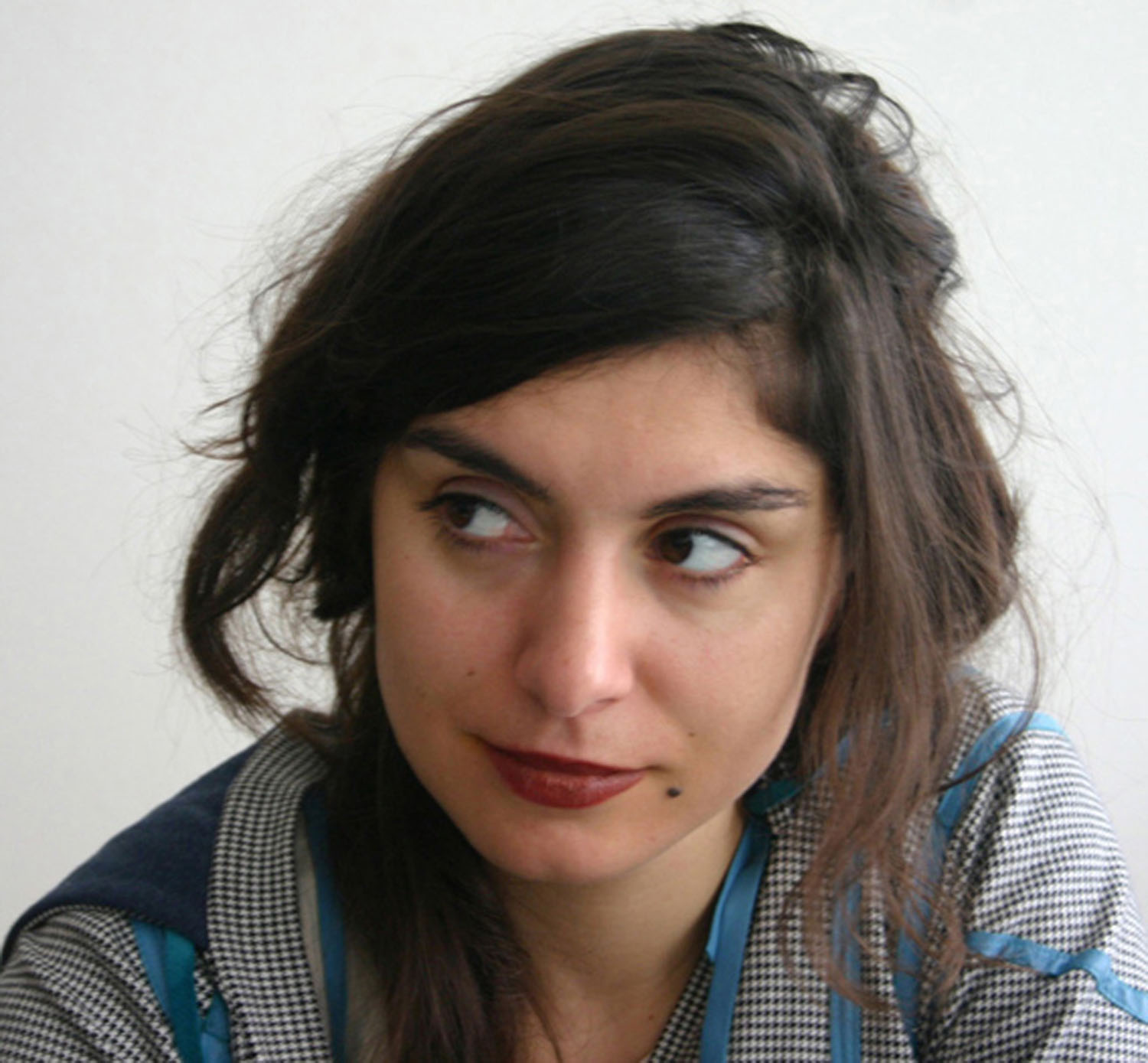 Woman with long, dark hair, a thoughtful expression, and red lips, wearing a checkered top.