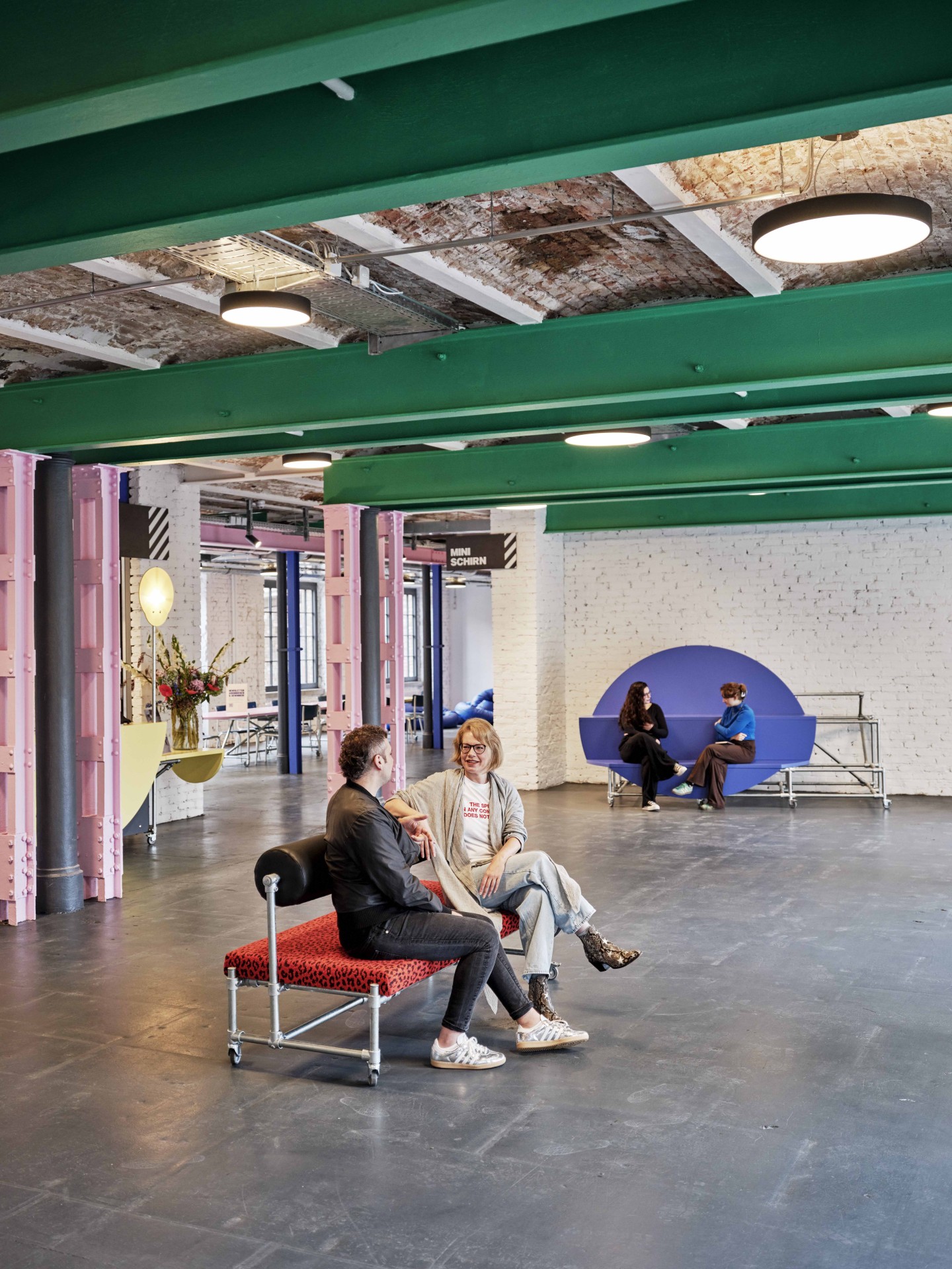 Zwei Personen sitzen auf einer Couch und unterhalten sich in einem modernen, hellen Raum mit bunten Akzenten.