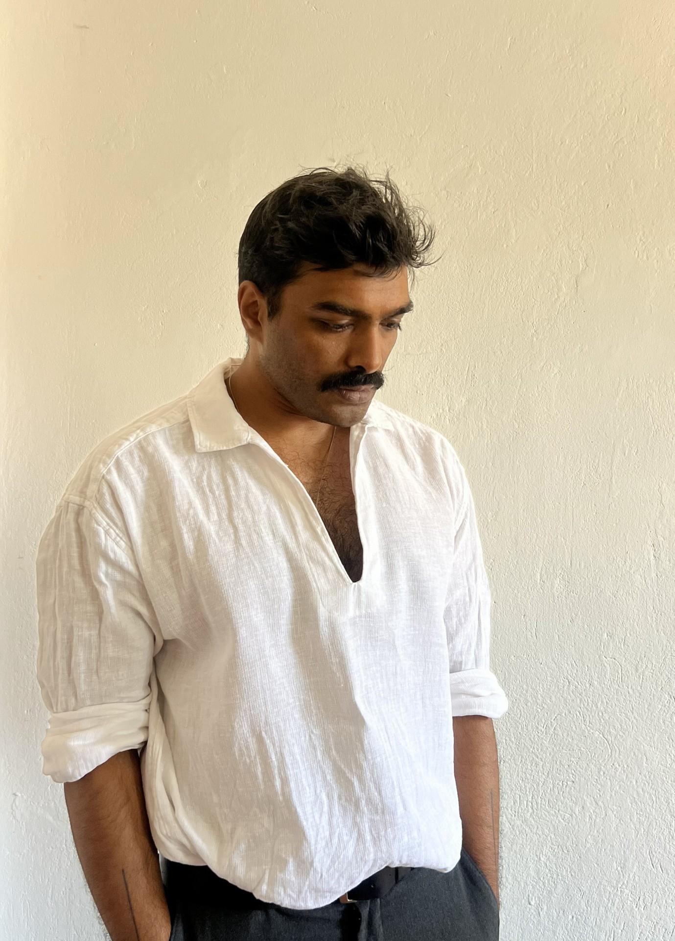 Mann in einem weißen Hemd steht vor einer neutralen Wand und schaut nachdenklich zu Boden.