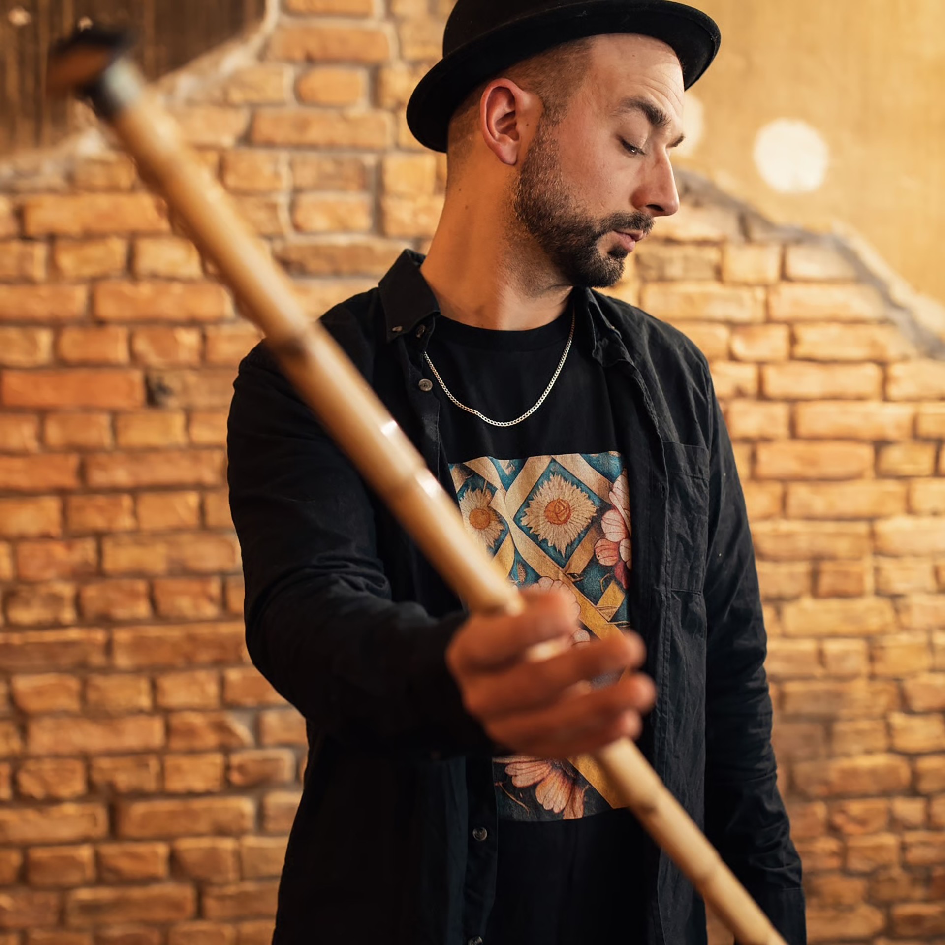 Mann mit Hut und Kette hält einen Bambusstab in einem Raum mit Backsteinmauer.