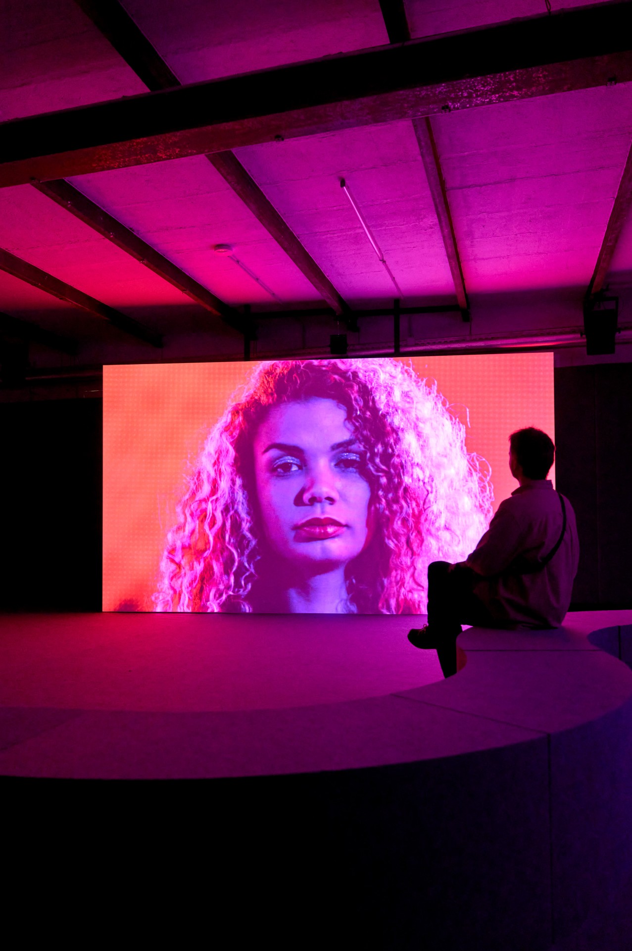 Eine Person sitzt vor einem großen, bunten Bildschirm, der ein Porträt in Pink- und Lilatönen zeigt.