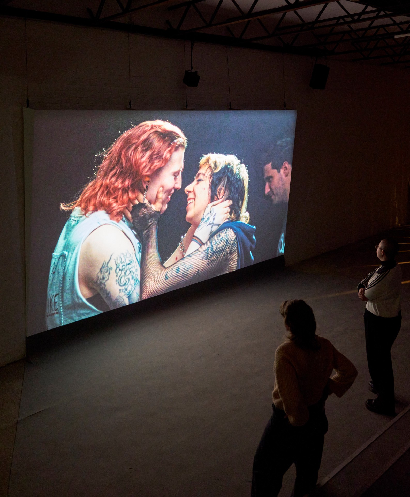 Two people kiss on a canvas while visitors observe the artwork in a room.