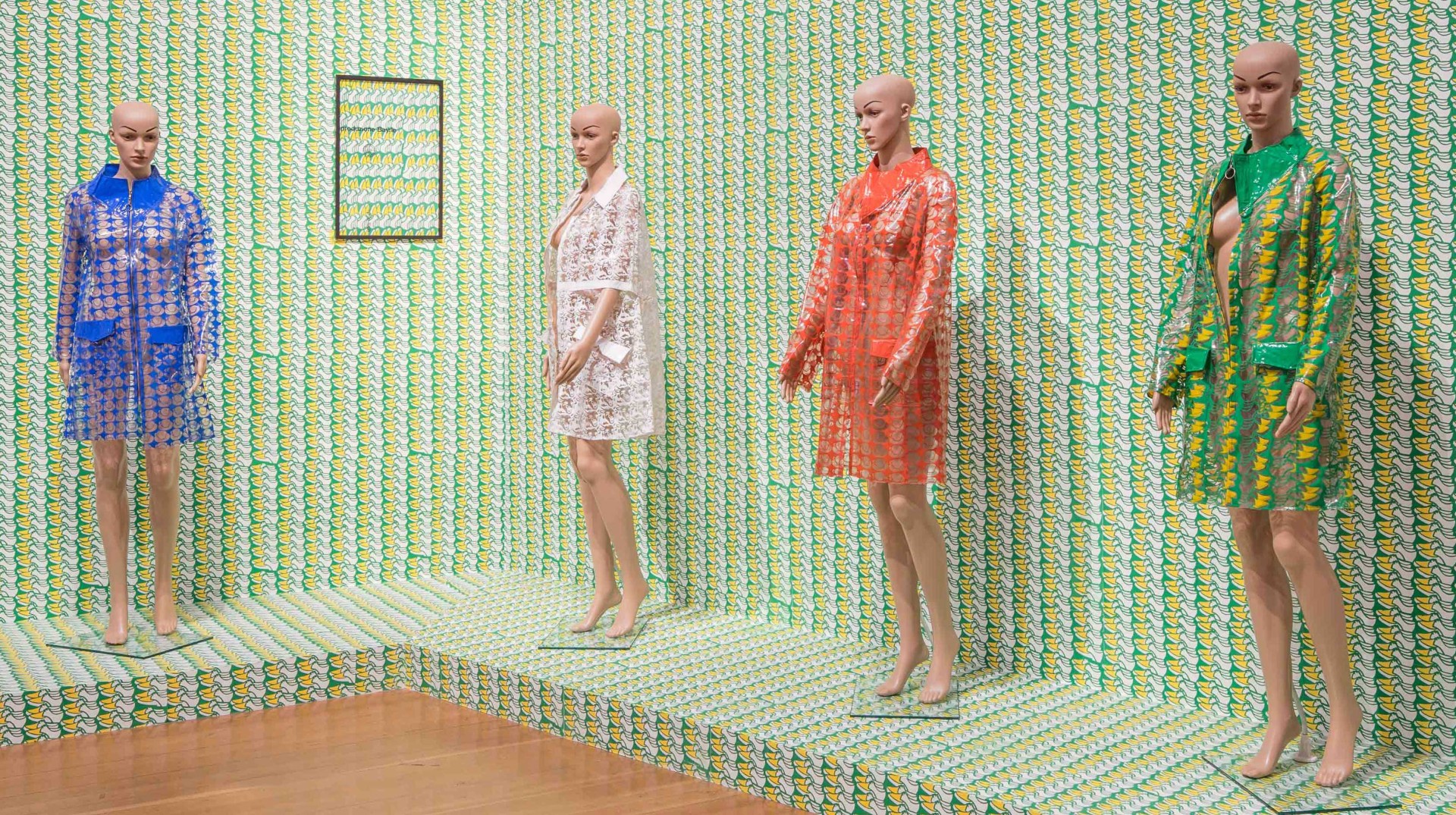 Vier Schaufensterpuppen in bunten, transparenten Mänteln vor einem dekorativen Hintergrund aus Musterpapier.