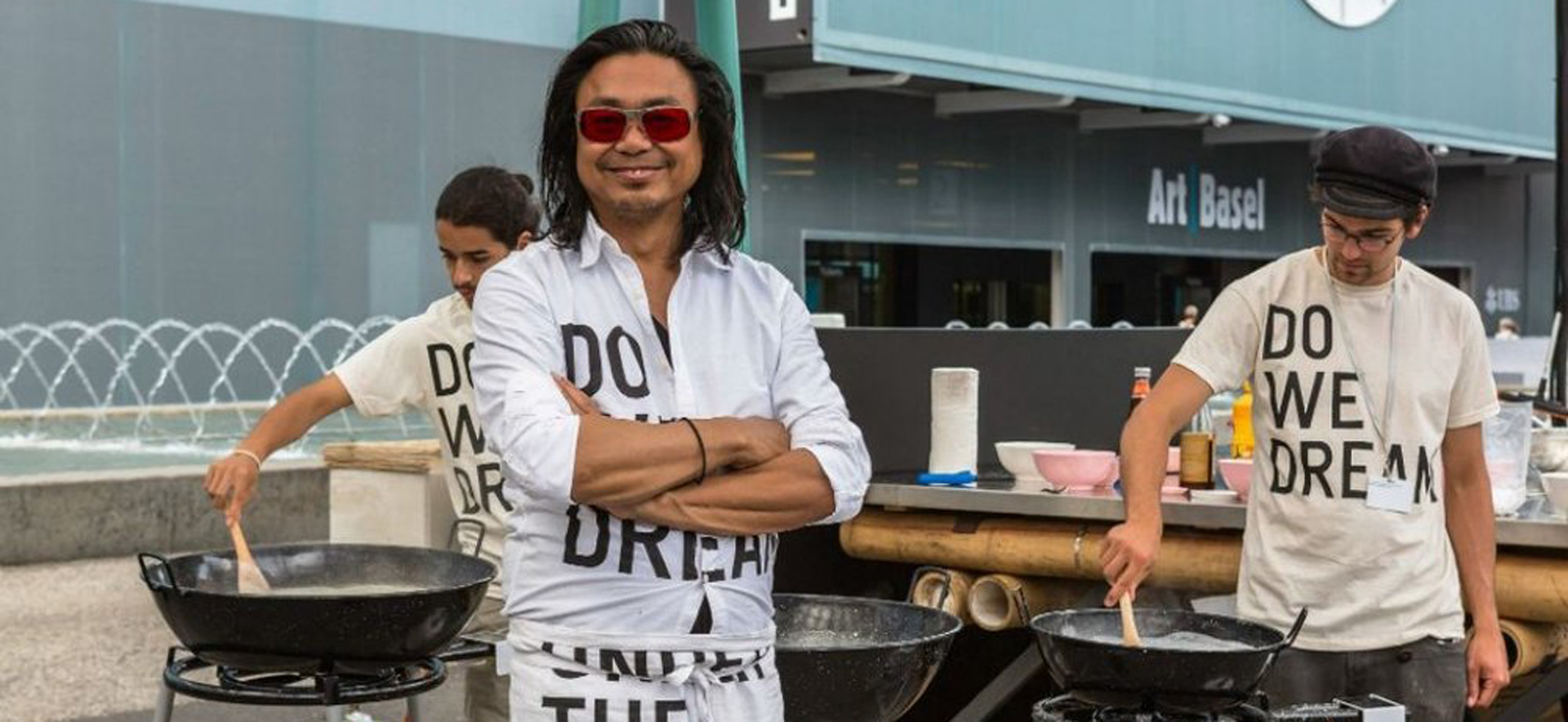 A person wearing sunglasses smiles in front of stoves while others prepare food in the background.