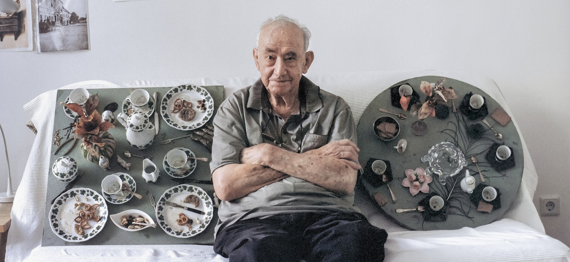 An older man sits on a sofa, surrounded by two coffee tables with dishes and decor.