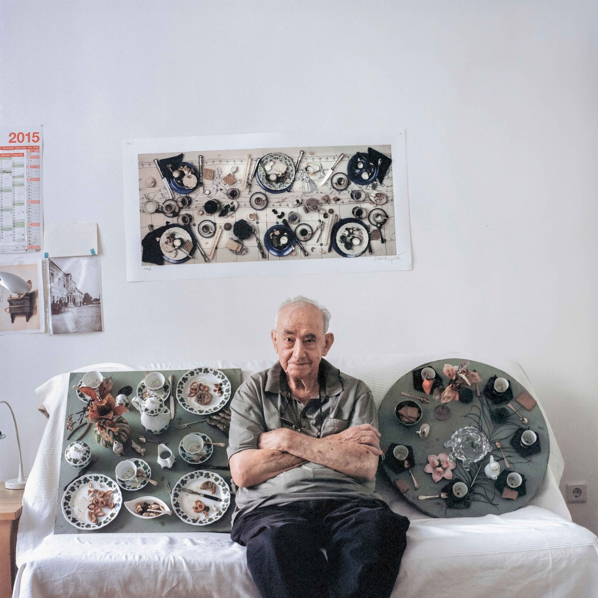An older man sits on a sofa, surrounded by dishes and a piece of artwork on the wall.