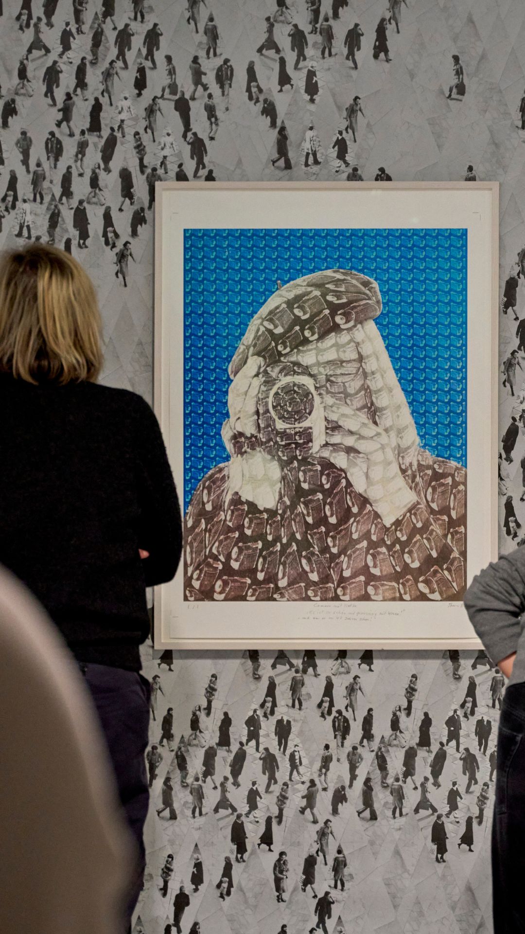 Two viewers are looking at an artwork depicting a folded person with a camera and a colorful background.