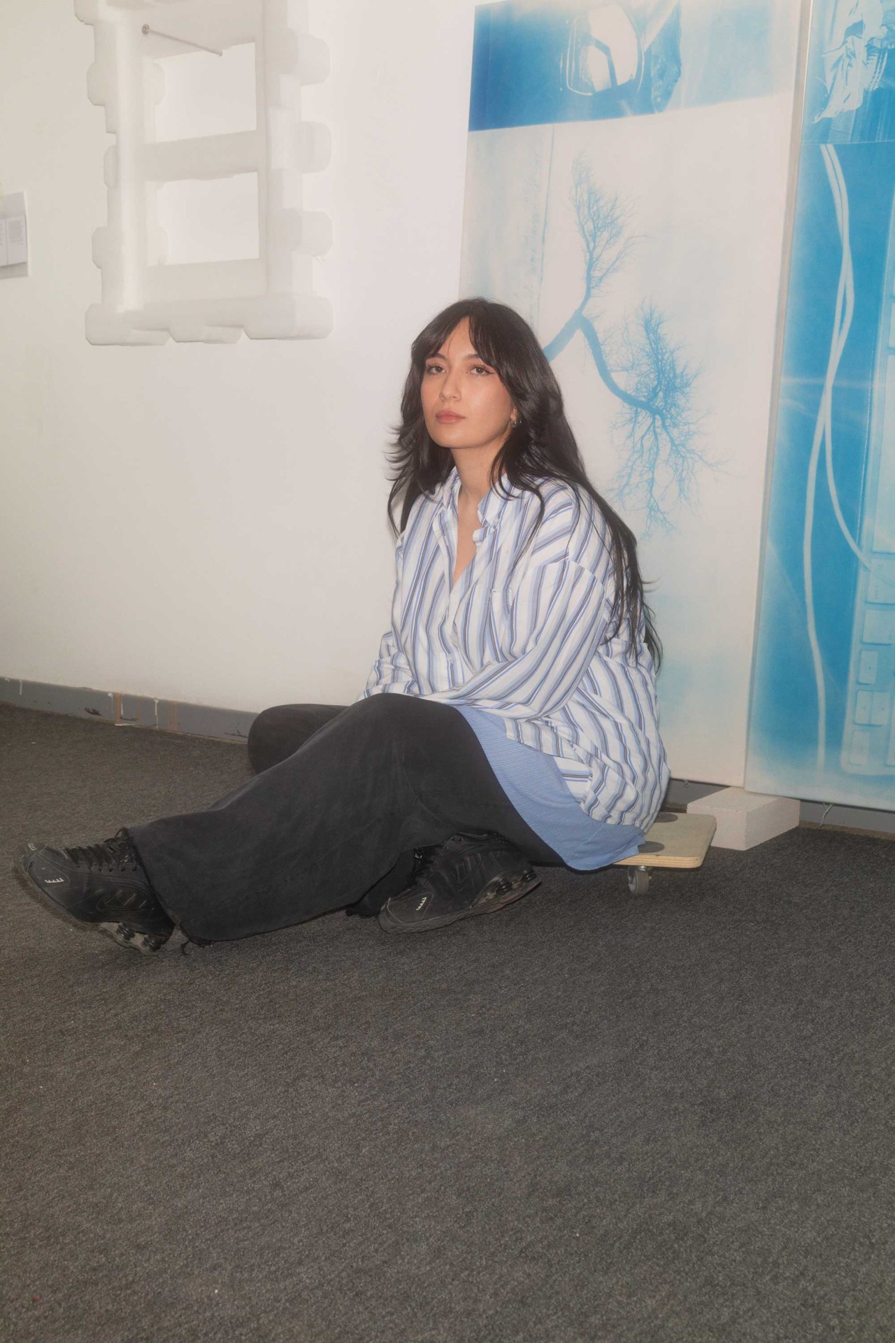 Frau sitzt auf dem Boden in einem Raum, trägt gestreifte Bluse und schwarze Hose, umgeben von Kunstwerken an der Wand.