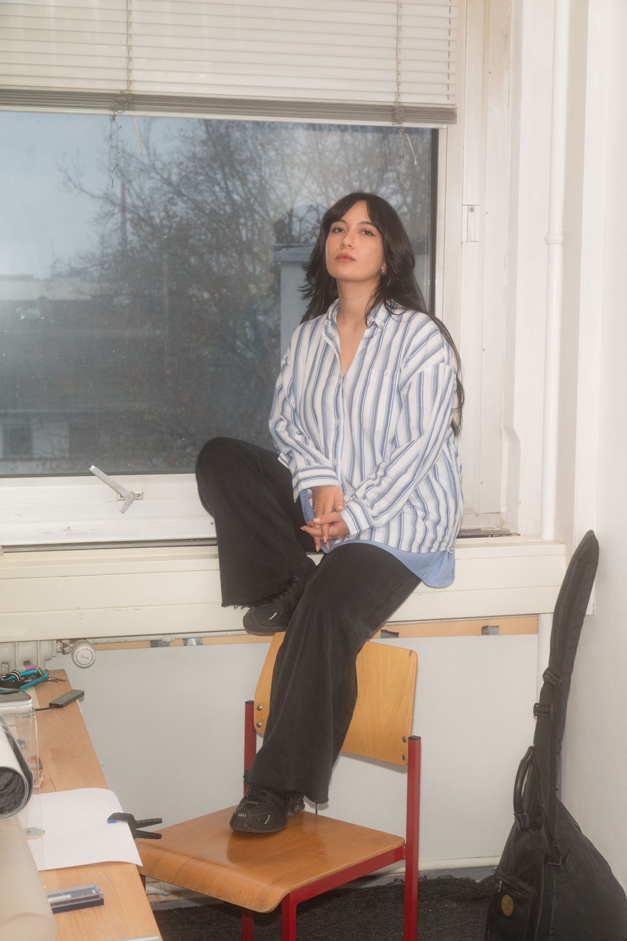 Frau mit langen Haaren sitzt auf dem Fensterbrett, trägt eine gestreifte Bluse und blickt nachdenklich hinaus.