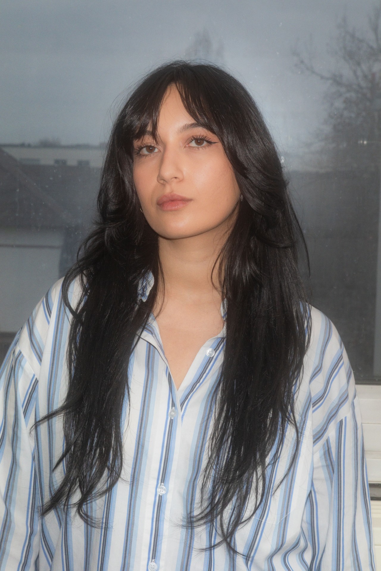 Junge Frau mit langen, dunklen Haaren und gestreiftem Hemd steht vor einem Fenster mit gedÀmpften LichtverhÀltnissen.