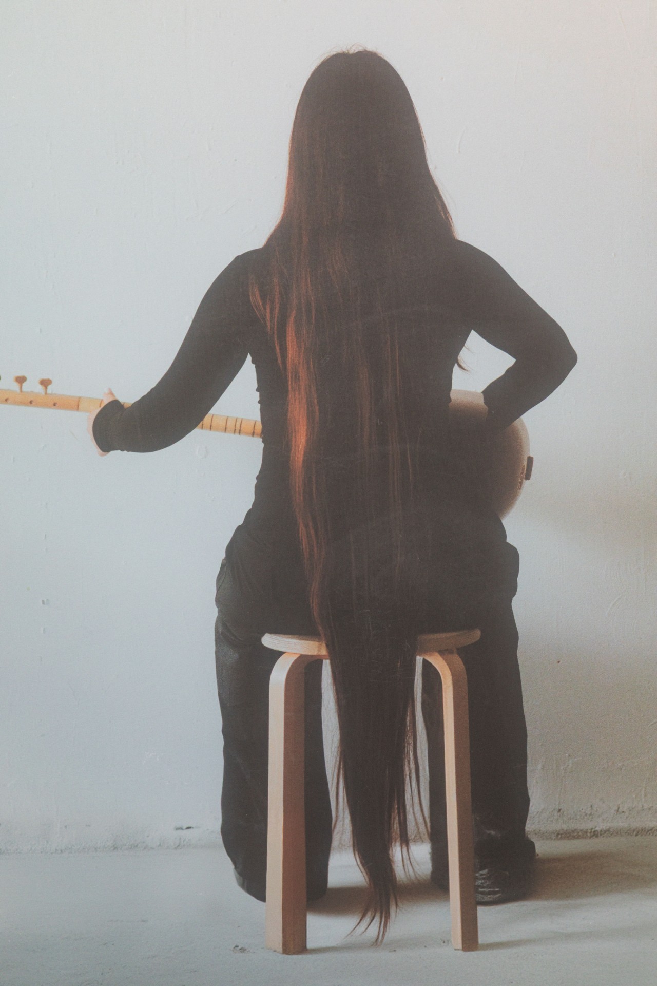 Person mit langen Haaren sitzt auf einem Hocker, hält ein Musikinstrument und sieht von hinten.
