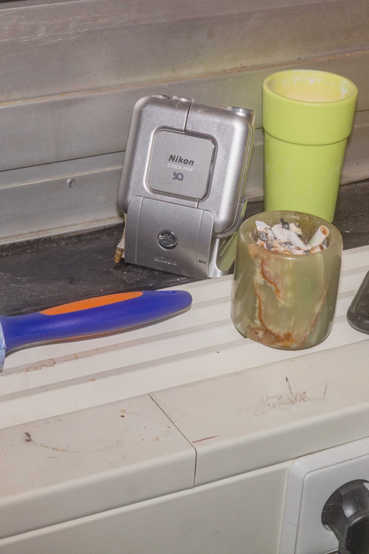 Eine Nikon Kamera, ein grüner Becher, ein Aschenbecher und ein blauer Werkzeuggriff auf einem Tisch.