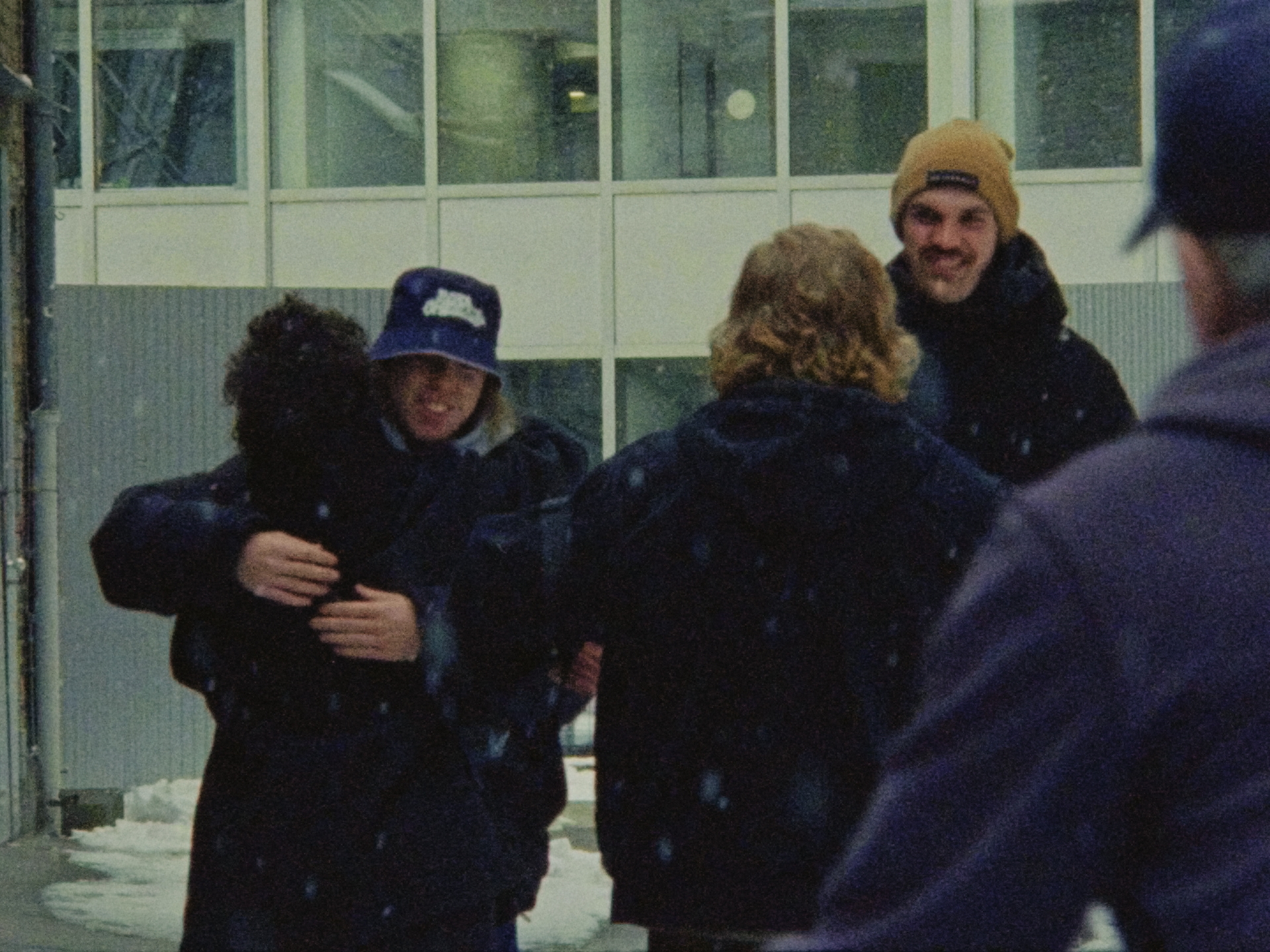 Gruppenzusammenhalt: Vier Personen umarmen sich in einer verschneiten Umgebung, Gebäude im Hintergrund. Freude und Freundschaft strahlen.