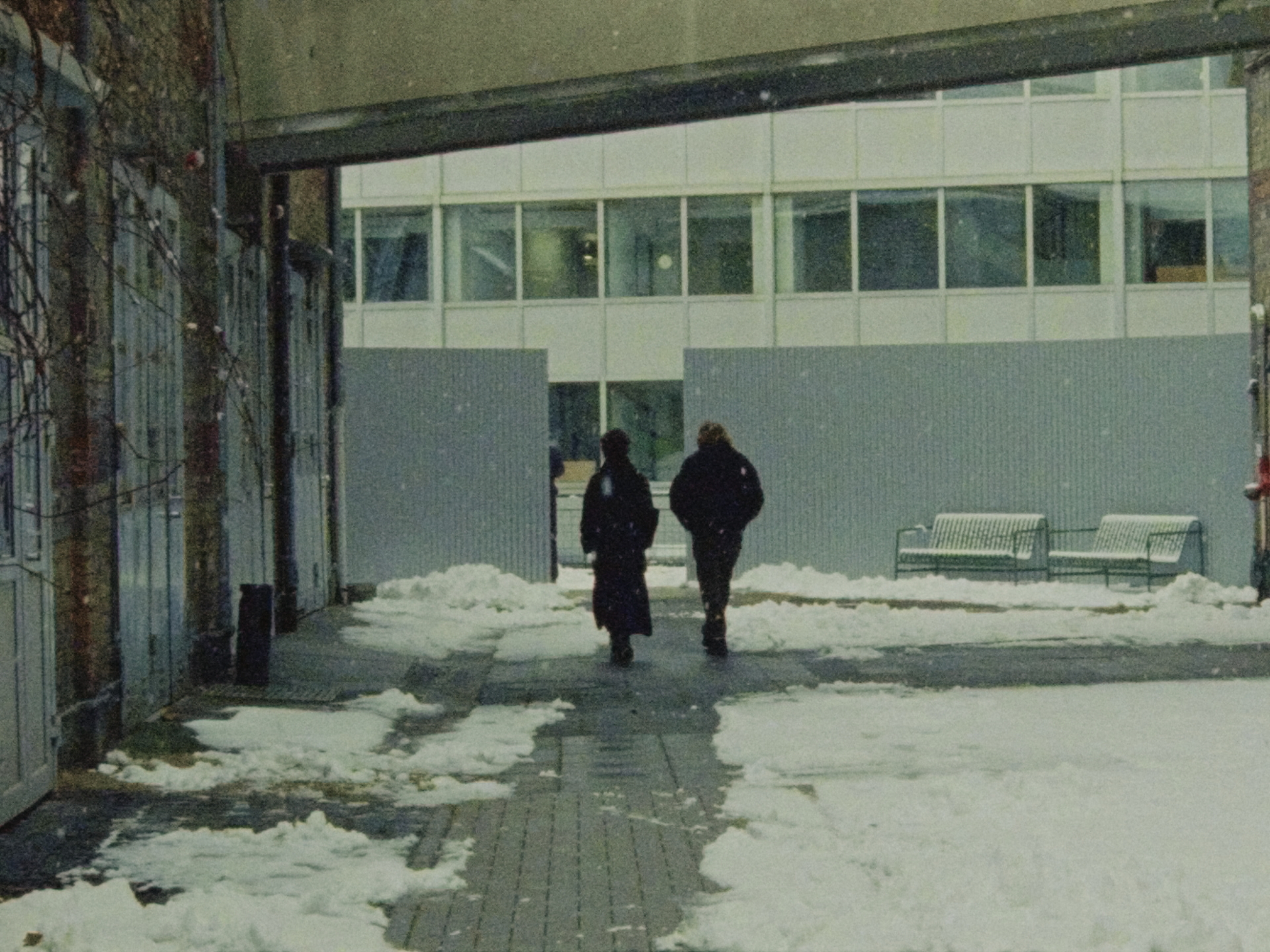 Zwei Personen gehen durch einen verschneiten Innenhof, umgeben von Gebäuden und einer grauen Mauer.