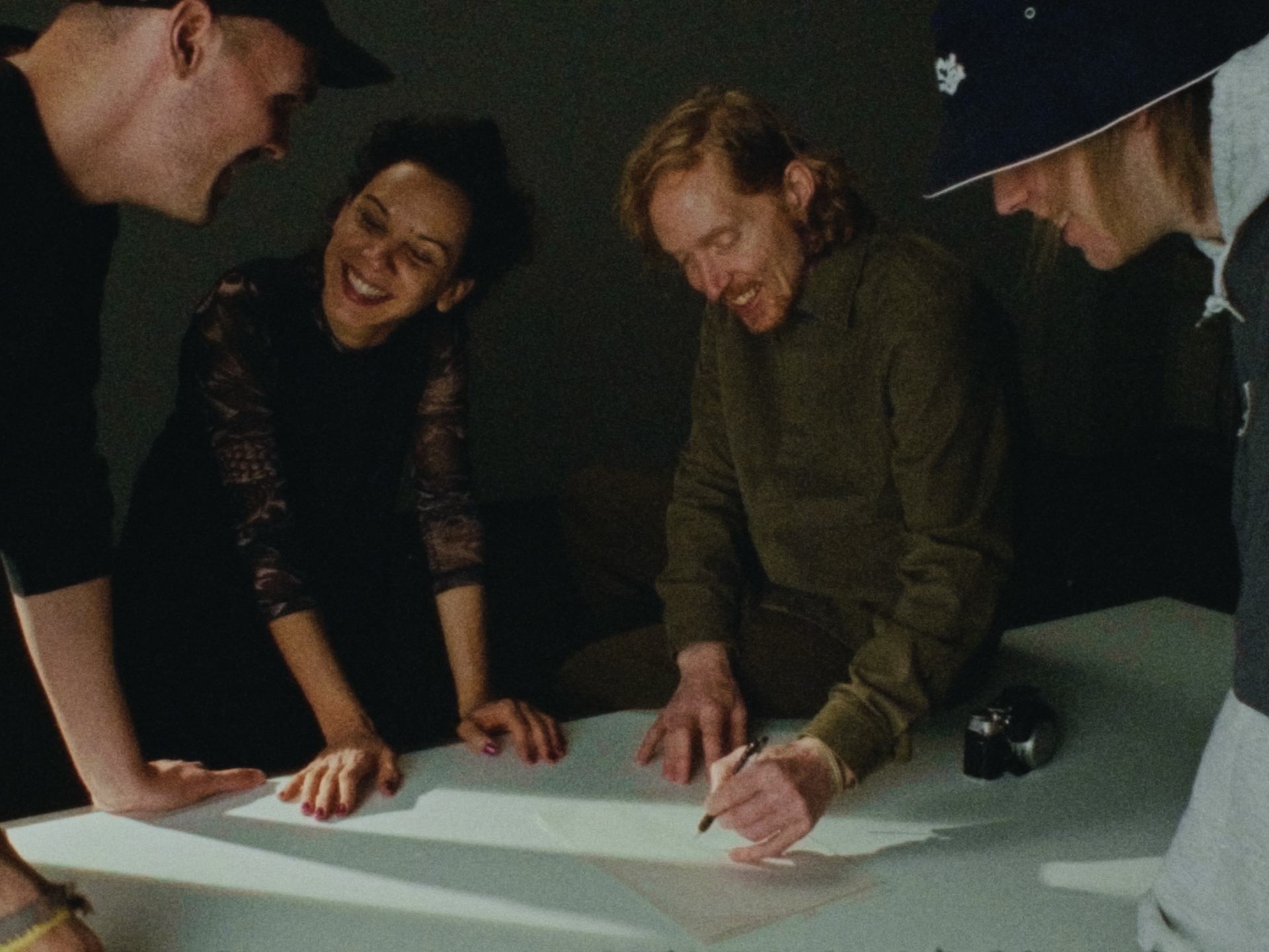 Vier Personen lachen und arbeiten gemeinsam an einem Tisch, während einer von ihnen in ein Notizbuch notiert.