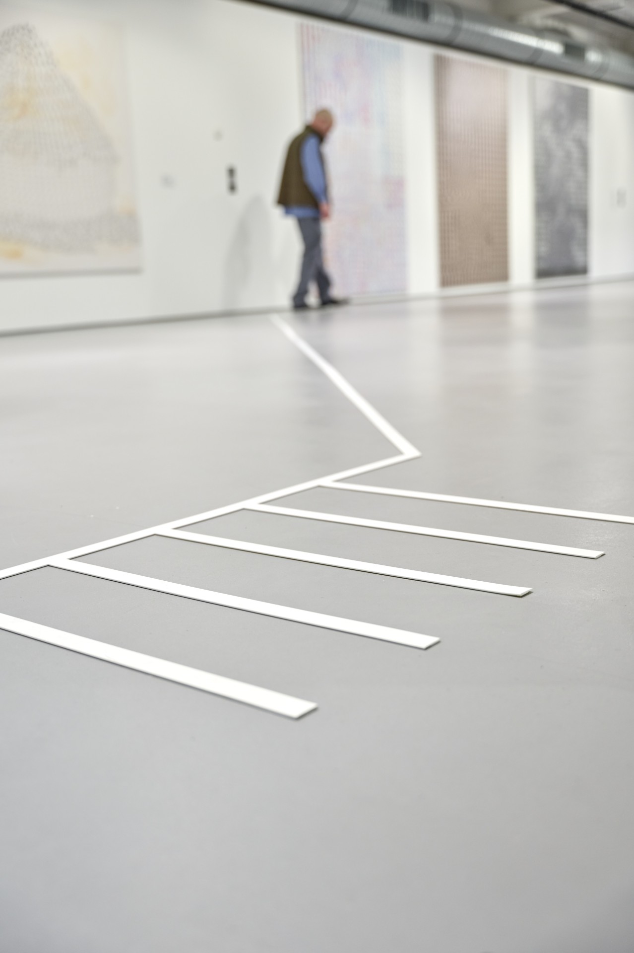 Eine Galerie mit einem Mann, der neben einer markierten Bodenlinie steht; abstrakte Kunstwerke sind im Hintergrund sichtbar.