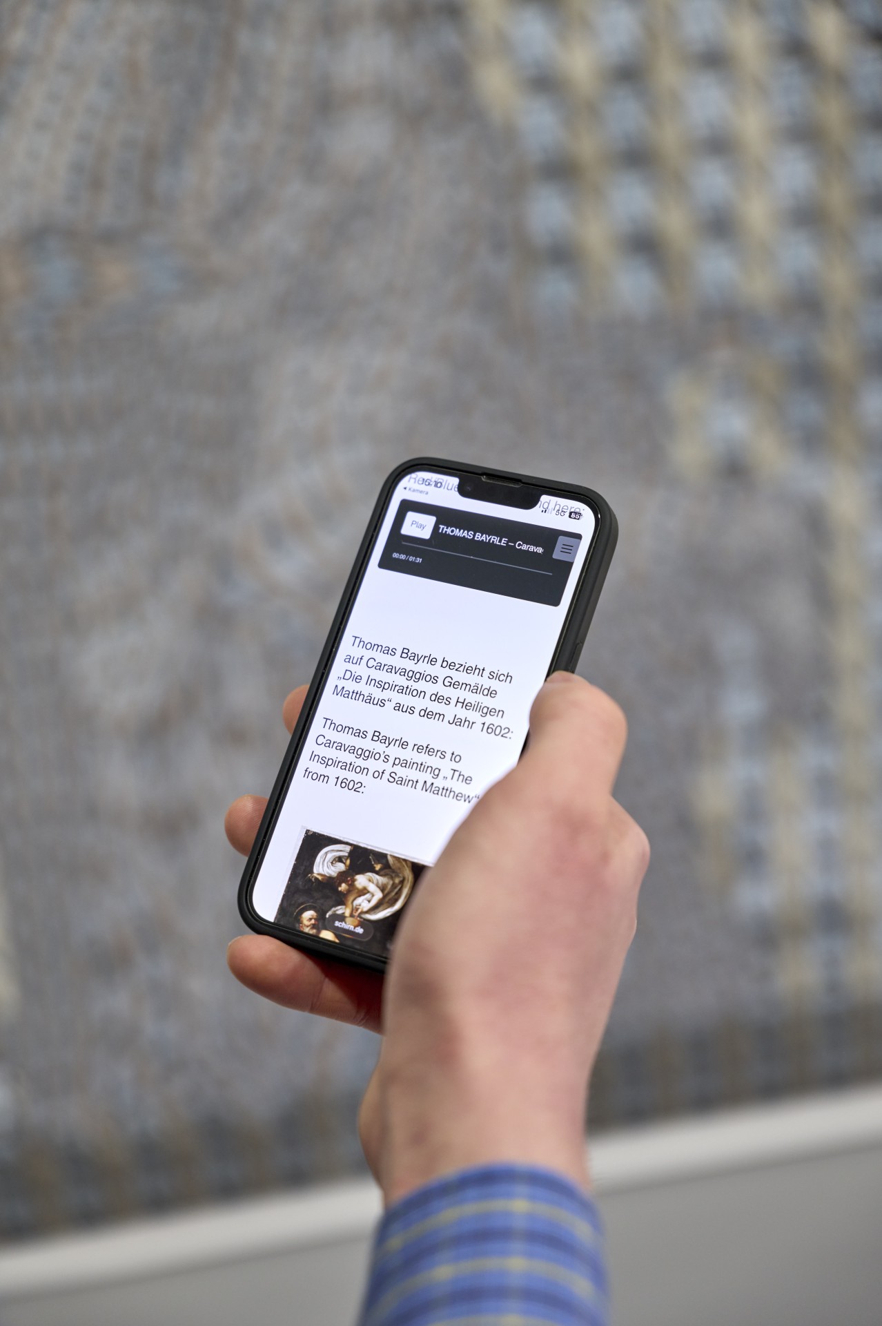 Person hält ein Smartphone mit Infos zu Thomas Bayrle und Caravaggios Gemälde „Die Inspiration des Heiligen Matthäus“.