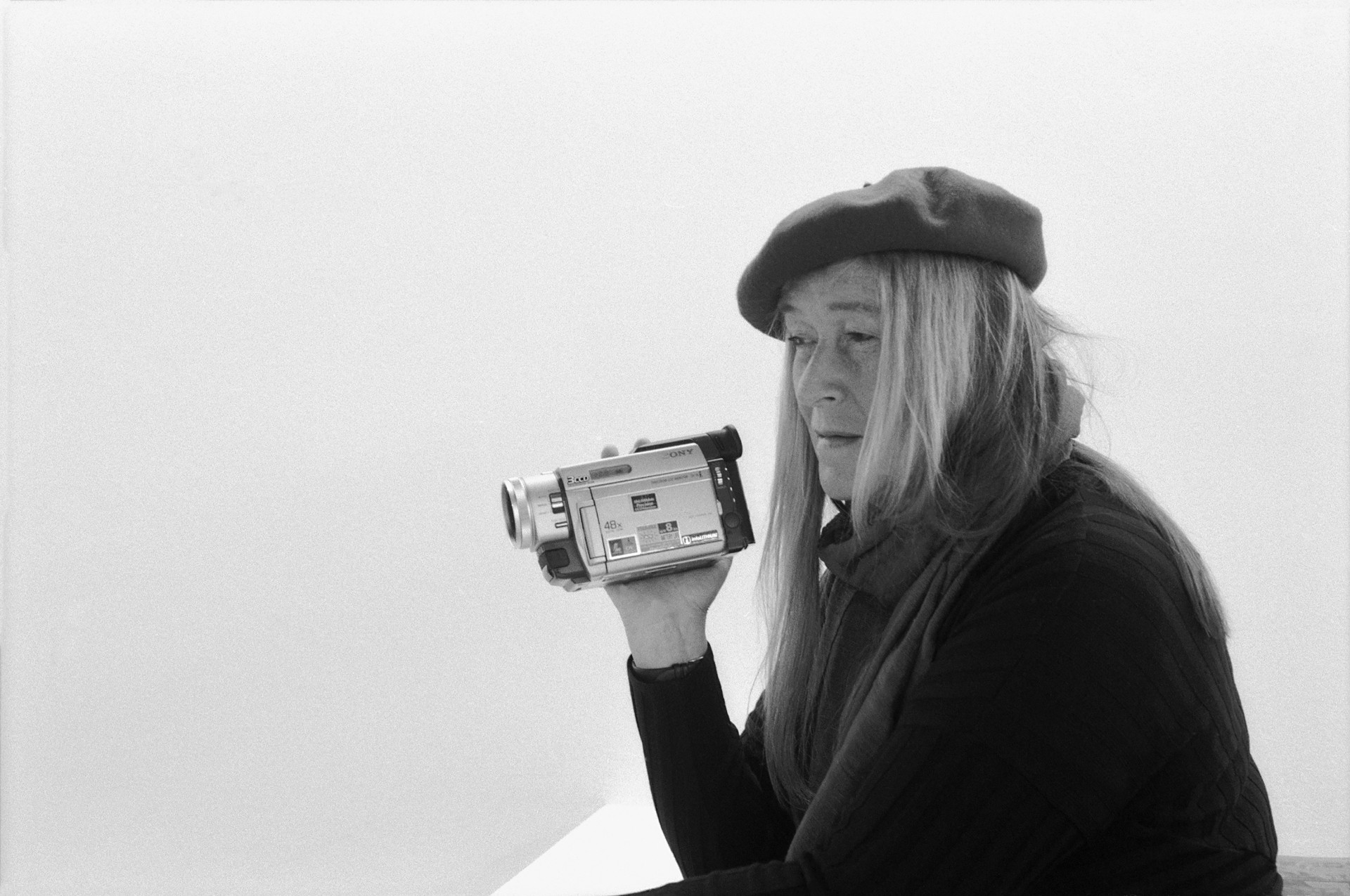 Frau mit langem, blondem Haar trägt eine Mütze und hält eine Videokamera, nachdenklicher Blick auf neutralem Hintergrund.