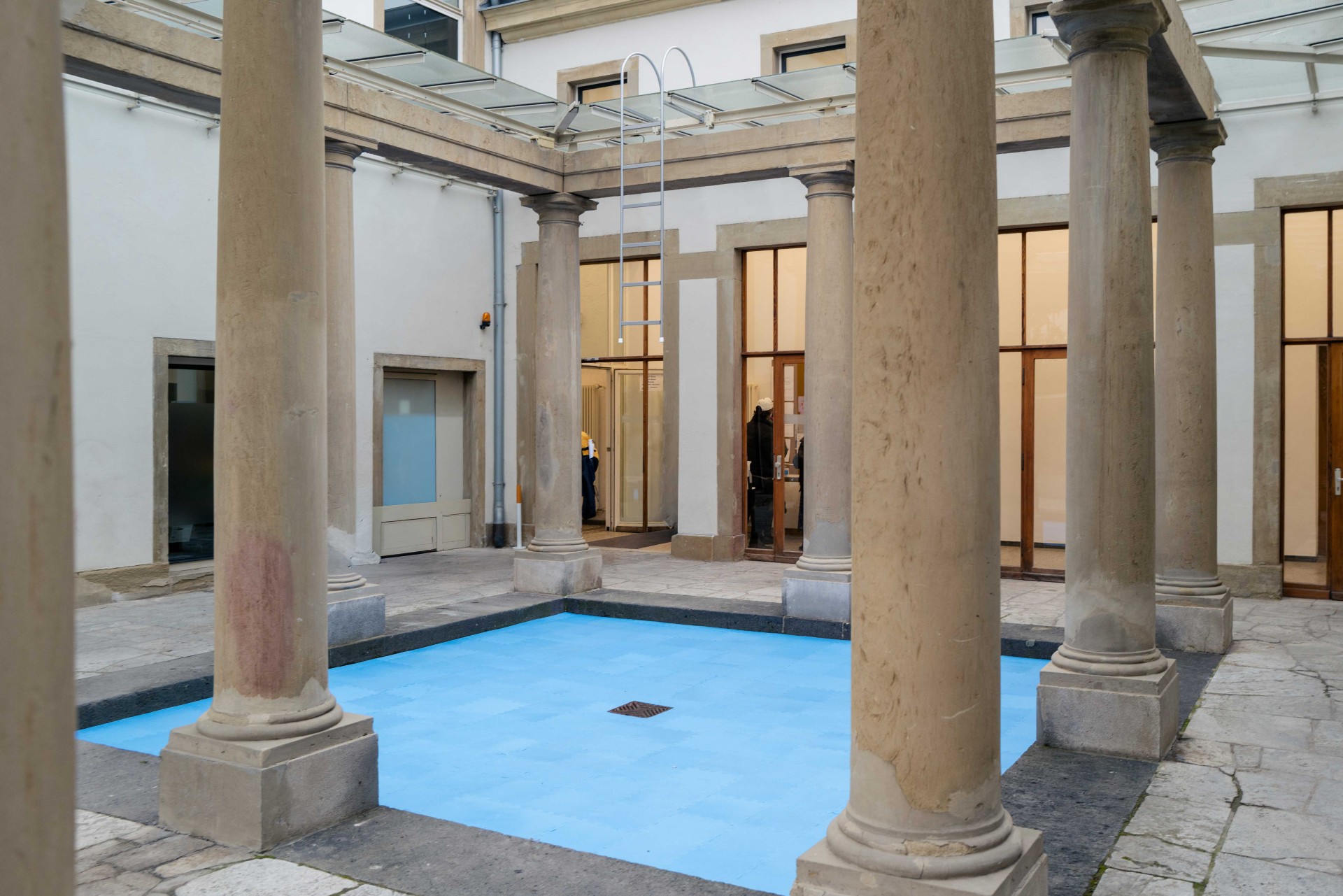 Innenhof mit Säulen und blauem Wasserbecken, umgeben von modernen Glas- und Holzelementen.
