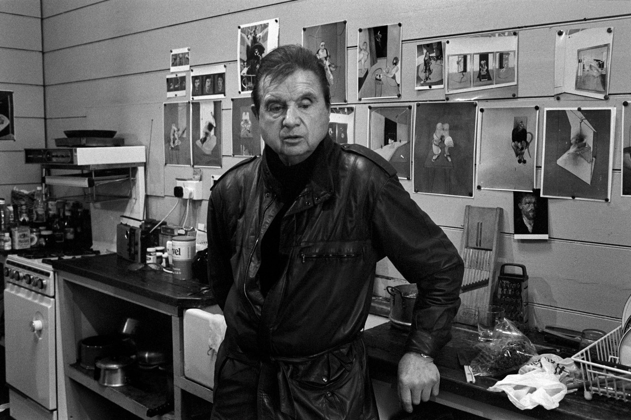 An older man in a leather jacket stands in a kitchen with photos on the wall, looking at the camera.