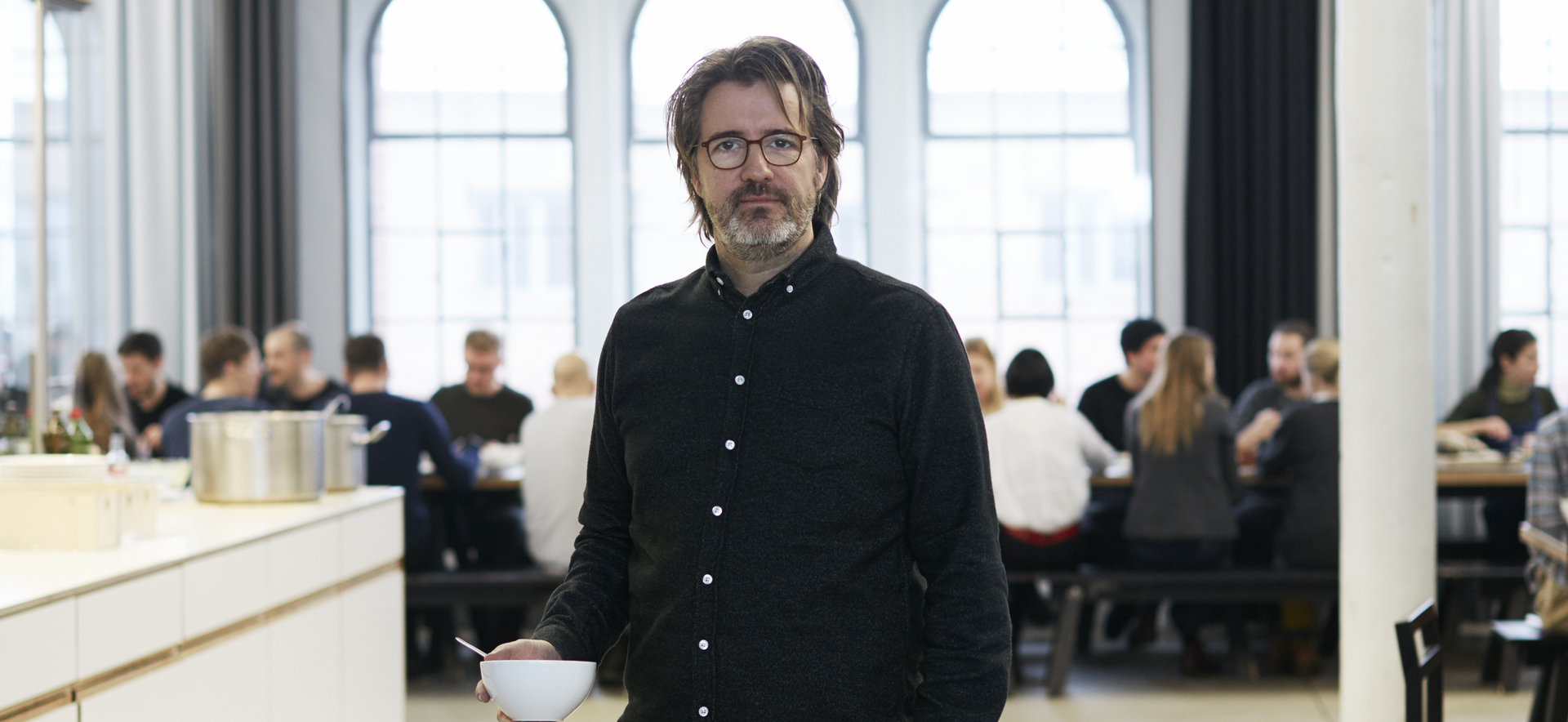 A man with glasses holds a bowl in a modern room, with people eating and chatting in the background.