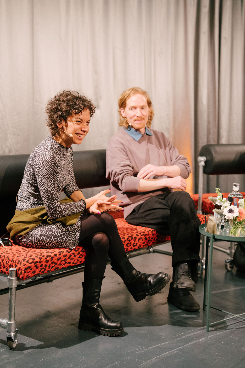 Two people sit on a sofa, talking to each other, surrounded by a modern setting and decorative elements.