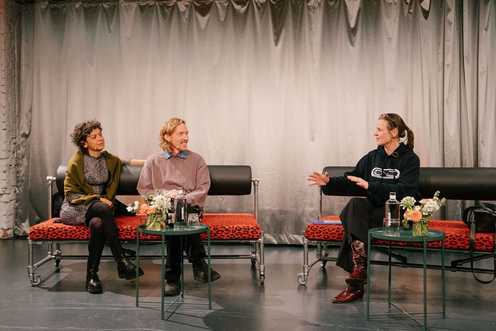 Three people sit on a stage, discussing and sharing ideas, surrounded by flowers and drinks.