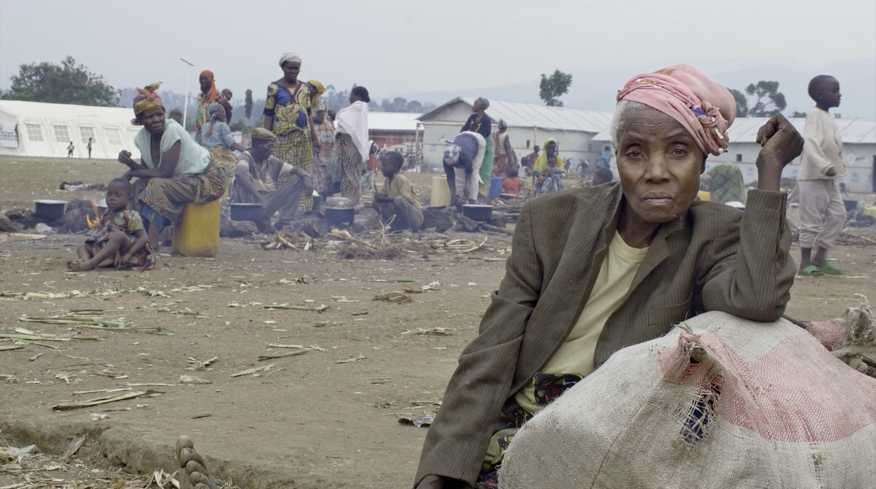 Eine ältere Frau sitzt nachdenklich auf einem Sack, umgeben von Menschen in einem belebten Lager.