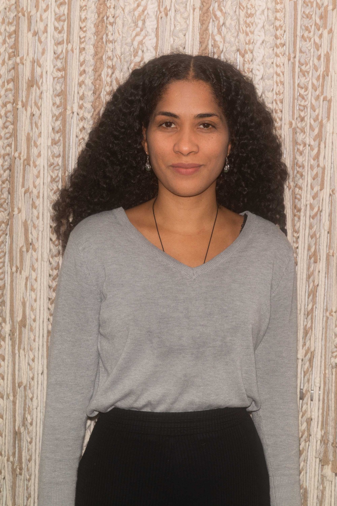 Frau mit lockigem Haar, grauem Pullover und schwarzer Hose vor einem bunten gemusterten Hintergrund.