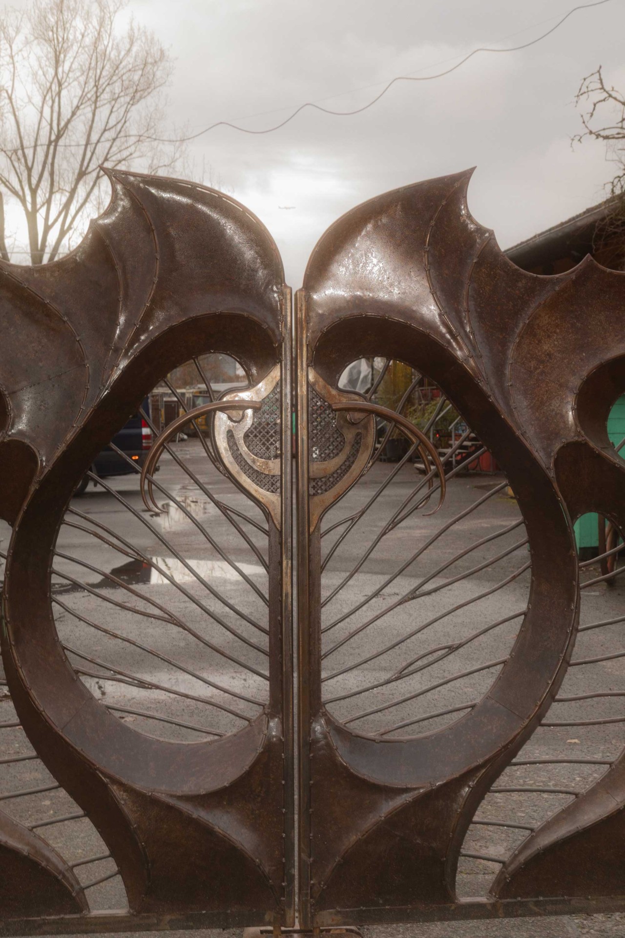 Verzierte Metalltür mit geschwungenem Design, die zu einem gepflasterten Hof unter bewölktem Himmel führt.