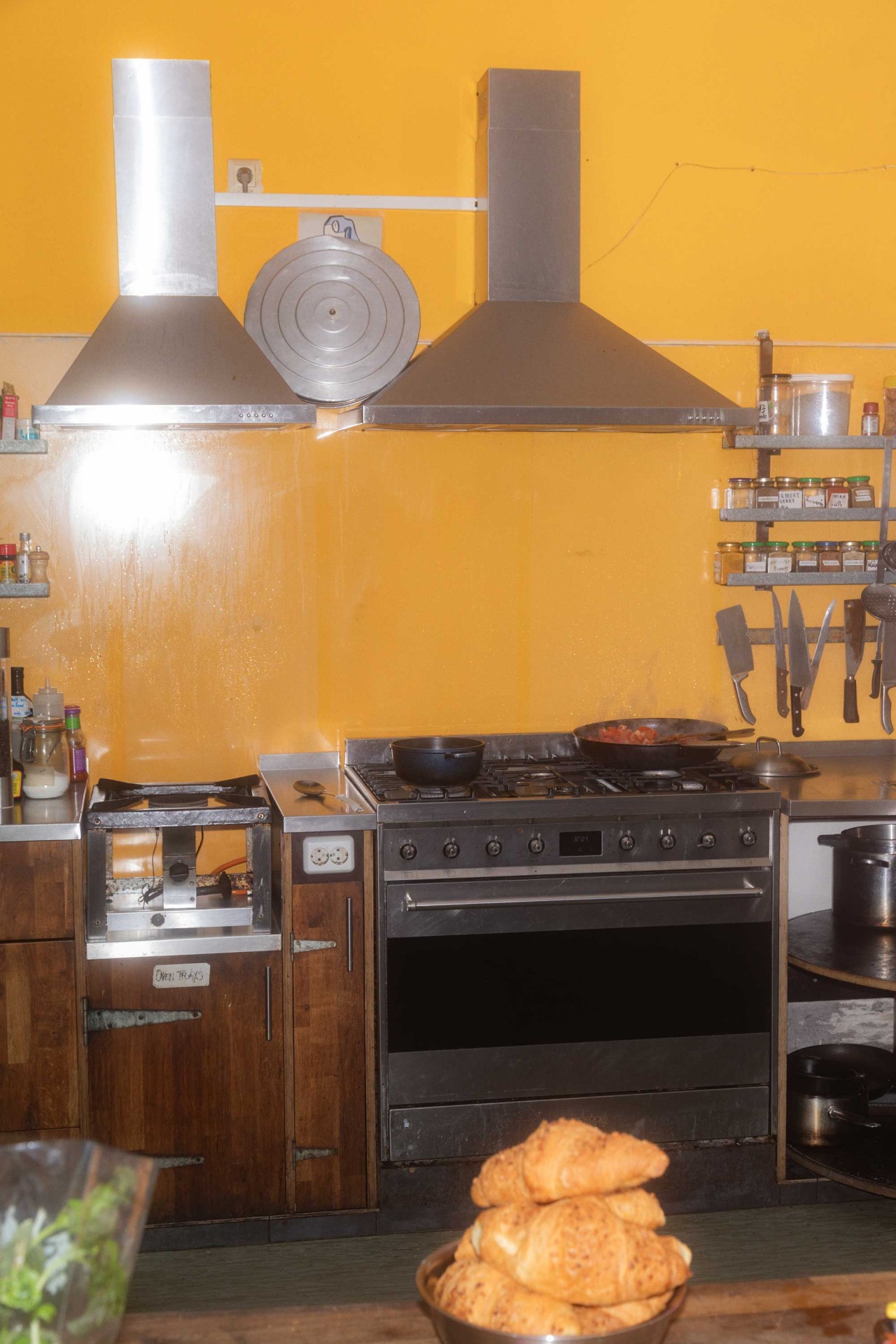 Küche mit gelber Wand, Edelstahlgeräten, Töpfen, Gewürzen und einem Stapel Croissants im Vordergrund.