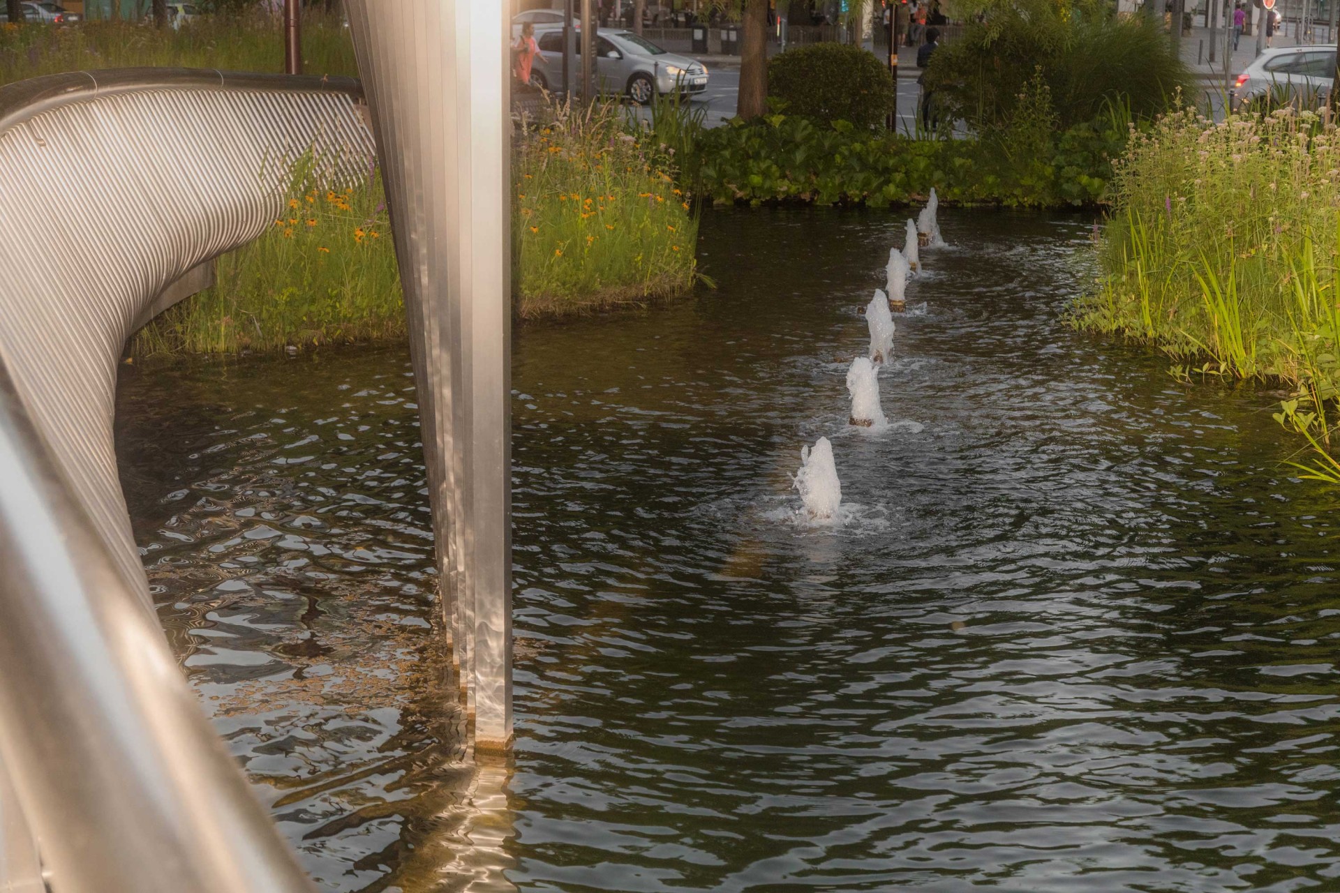 Moderne Brücke über ein Gewässer mit kleinen Springbrunnen und üppigem Grün am Ufer.