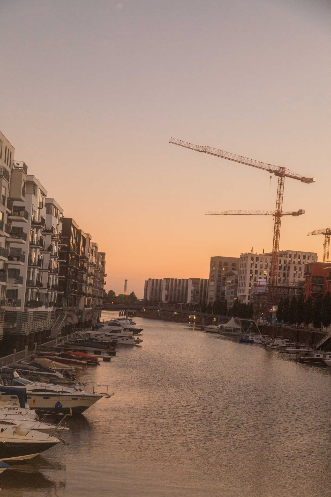 Blick auf einen Kanal mit Booten, modernen Gebäuden und Kränen während des Sonnenuntergangs.